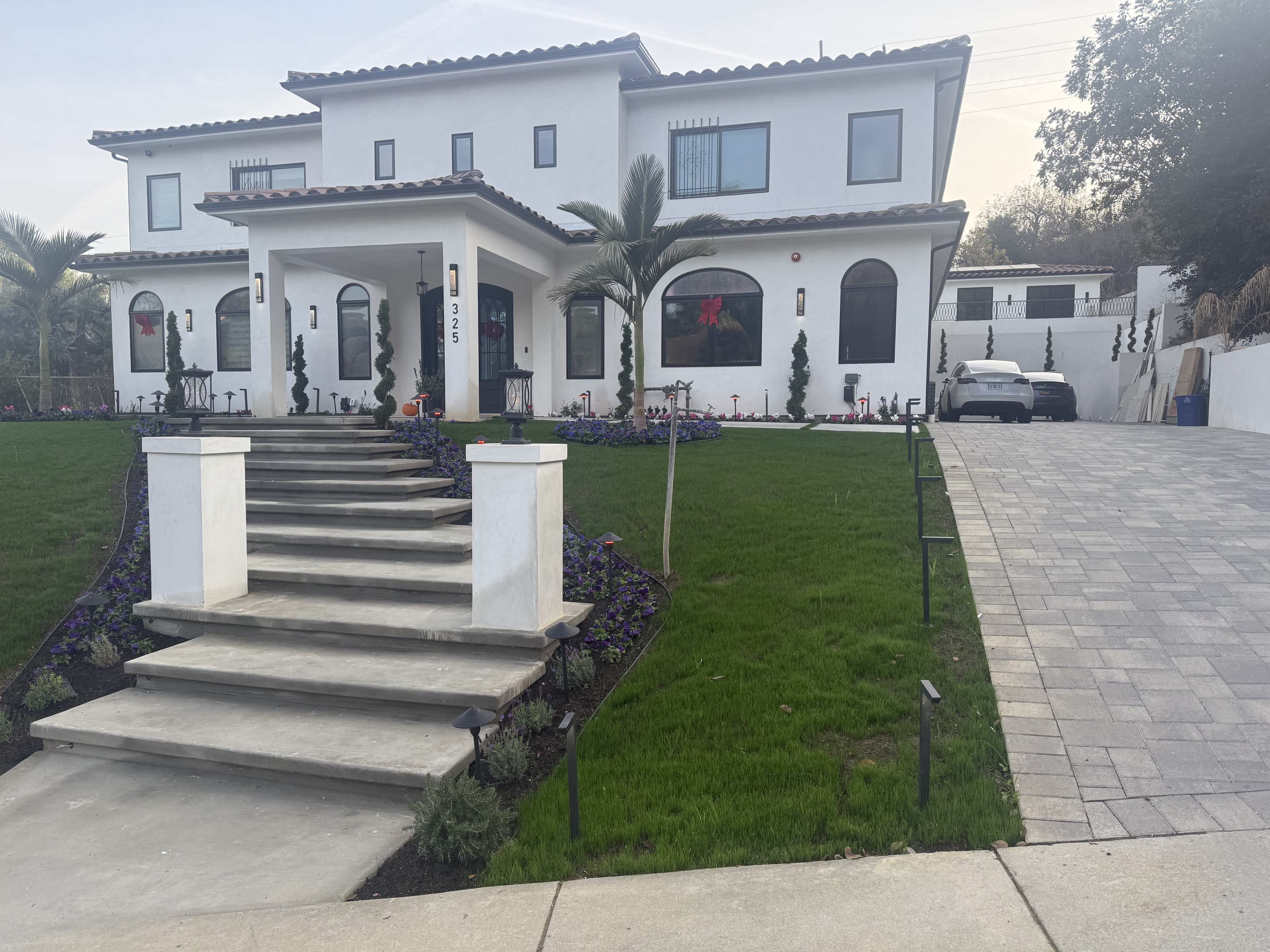 A large, modern house with a manicured lawn and stone steps leads up to the entrance, while a paved driveway extends to the side.