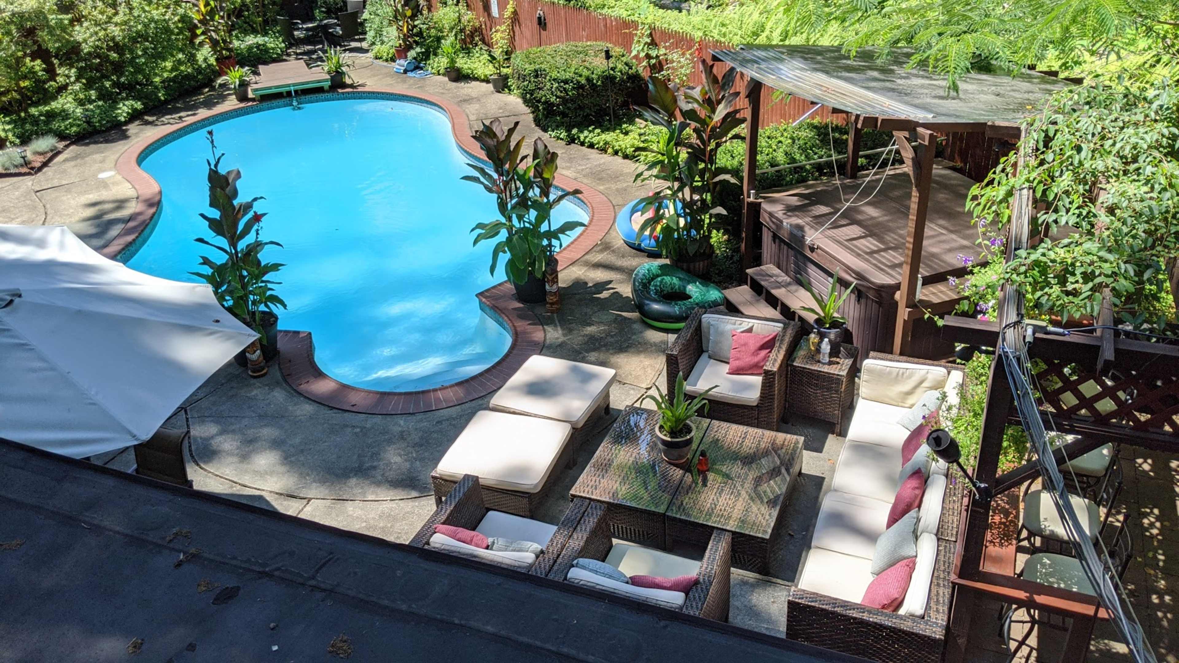 A landscaped backyard featuring a blue swimming pool surrounded by lounge chairs and potted plants, with a wooden deck and gazebo in the background.