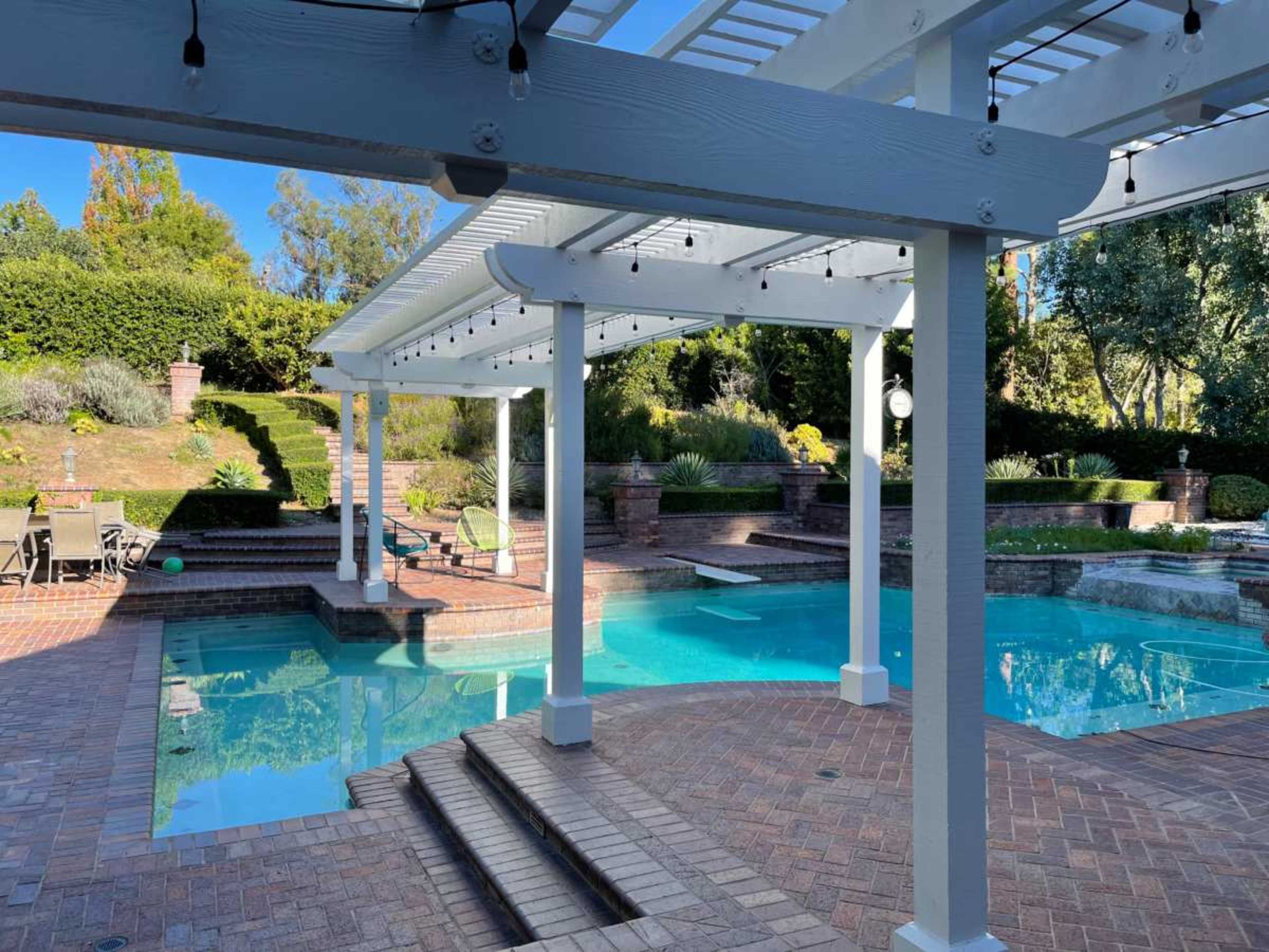 The image shows a spacious pool area surrounded by brick paving, with a covered patio and landscaped greenery in the background.