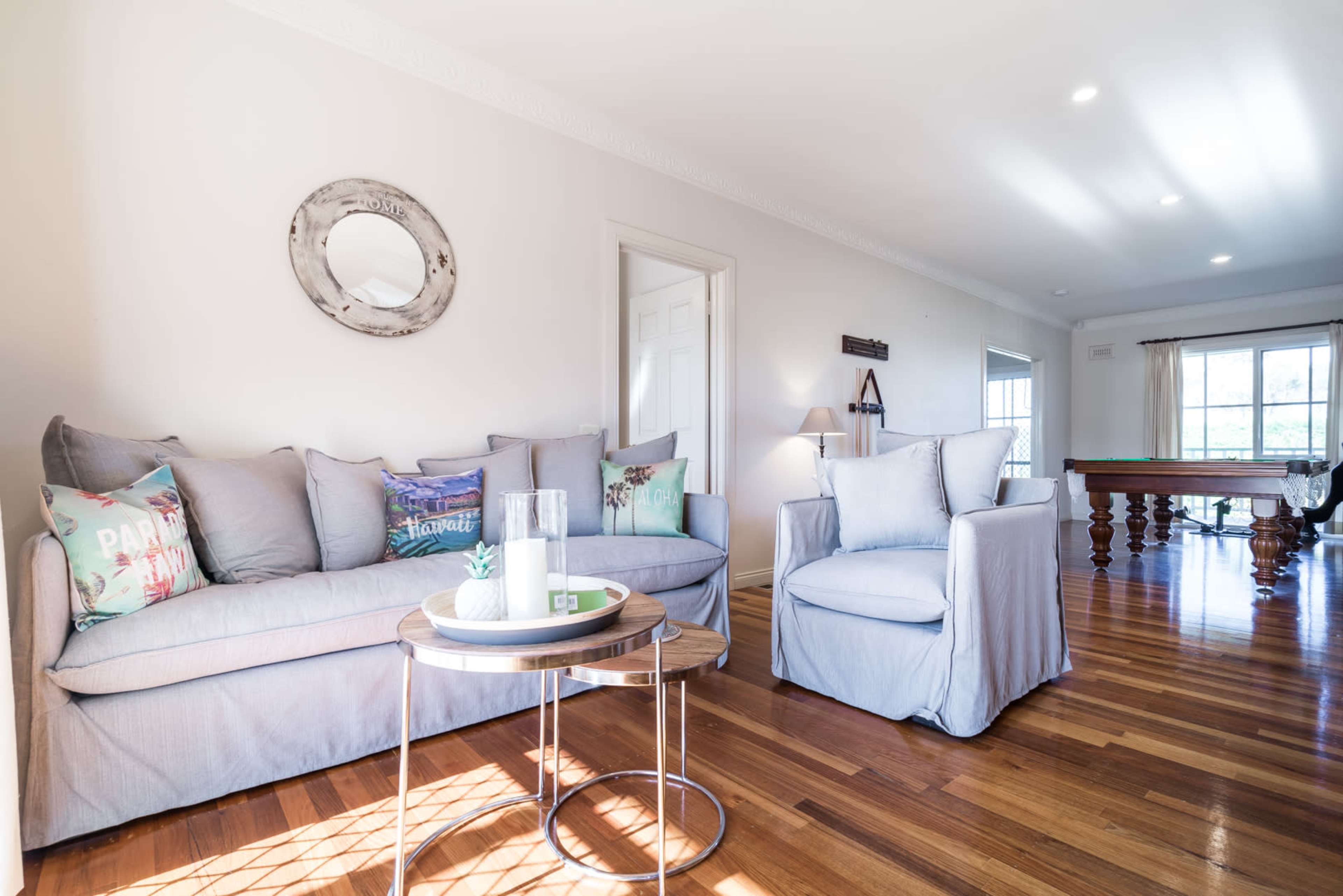 A cozy living room features a light gray sofa and armchair, a round table, and a pool table visible in the background.