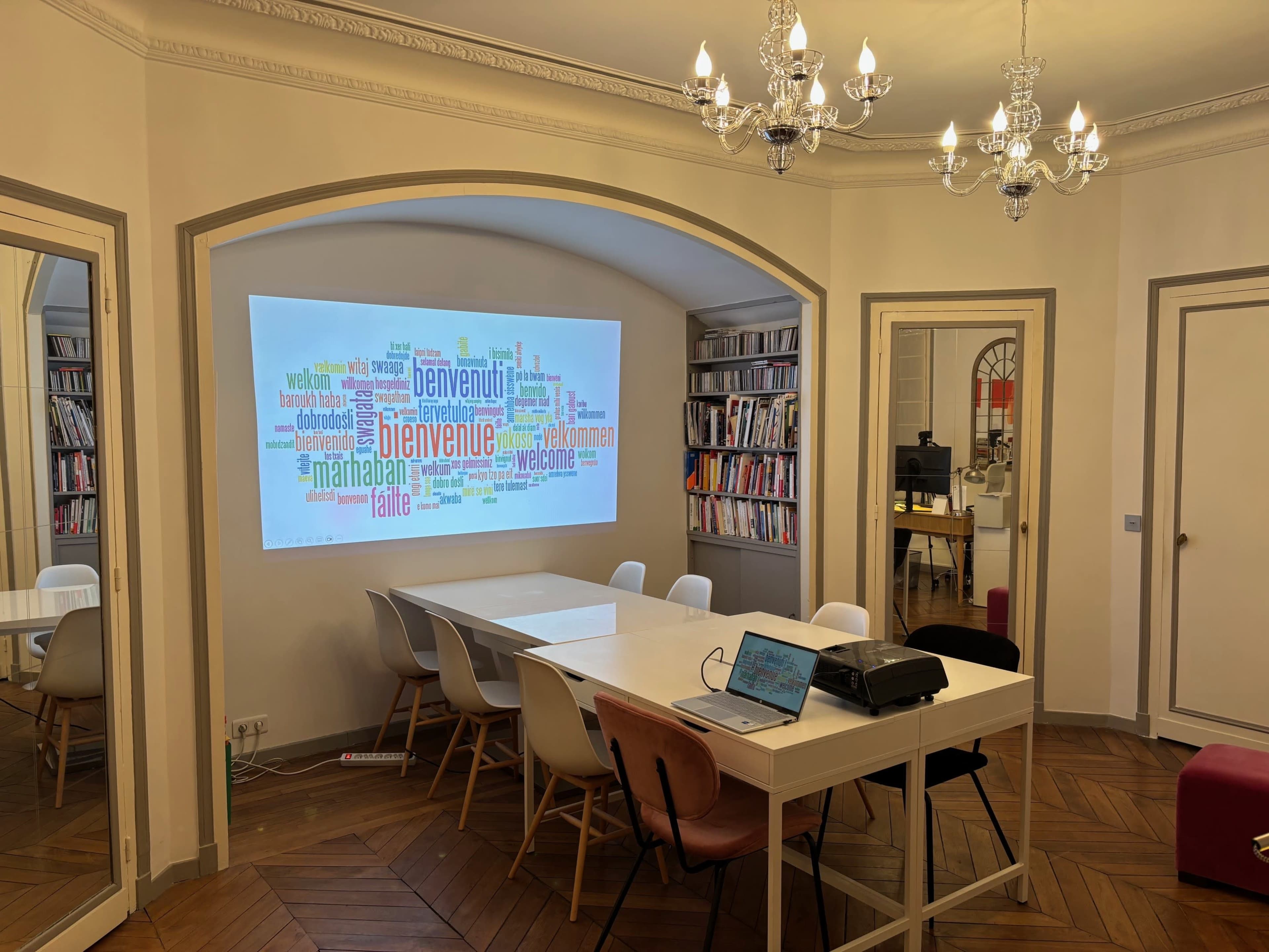 The image shows a modern conference room with a large table, several chairs, and a projection screen displaying a colorful word cloud, surrounded by bookshelves and decorative lighting.