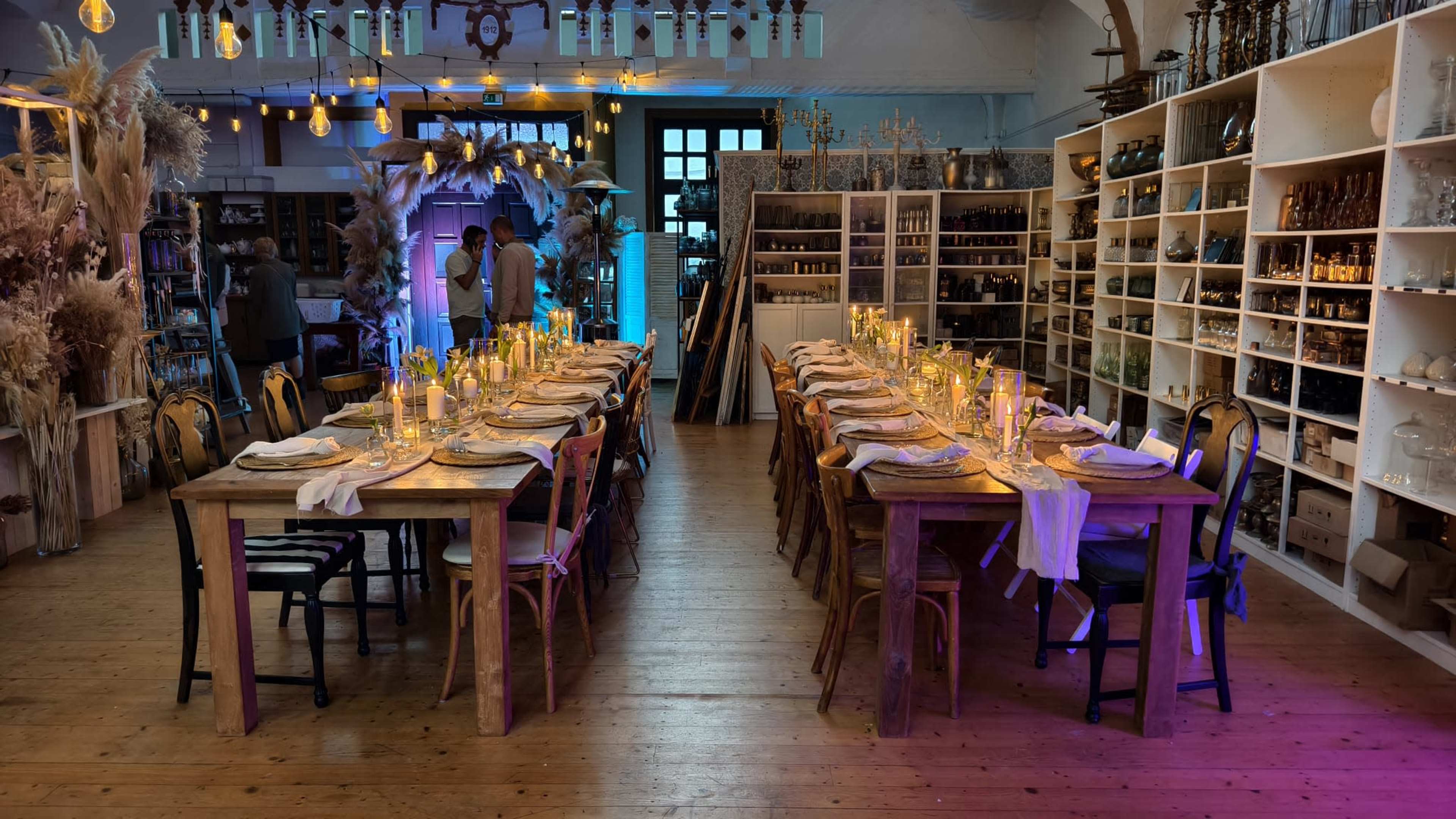 The image shows a spacious dining area with two long wooden tables set for a meal, adorned with candles and tableware, surrounded by shelves displaying various decorative items.