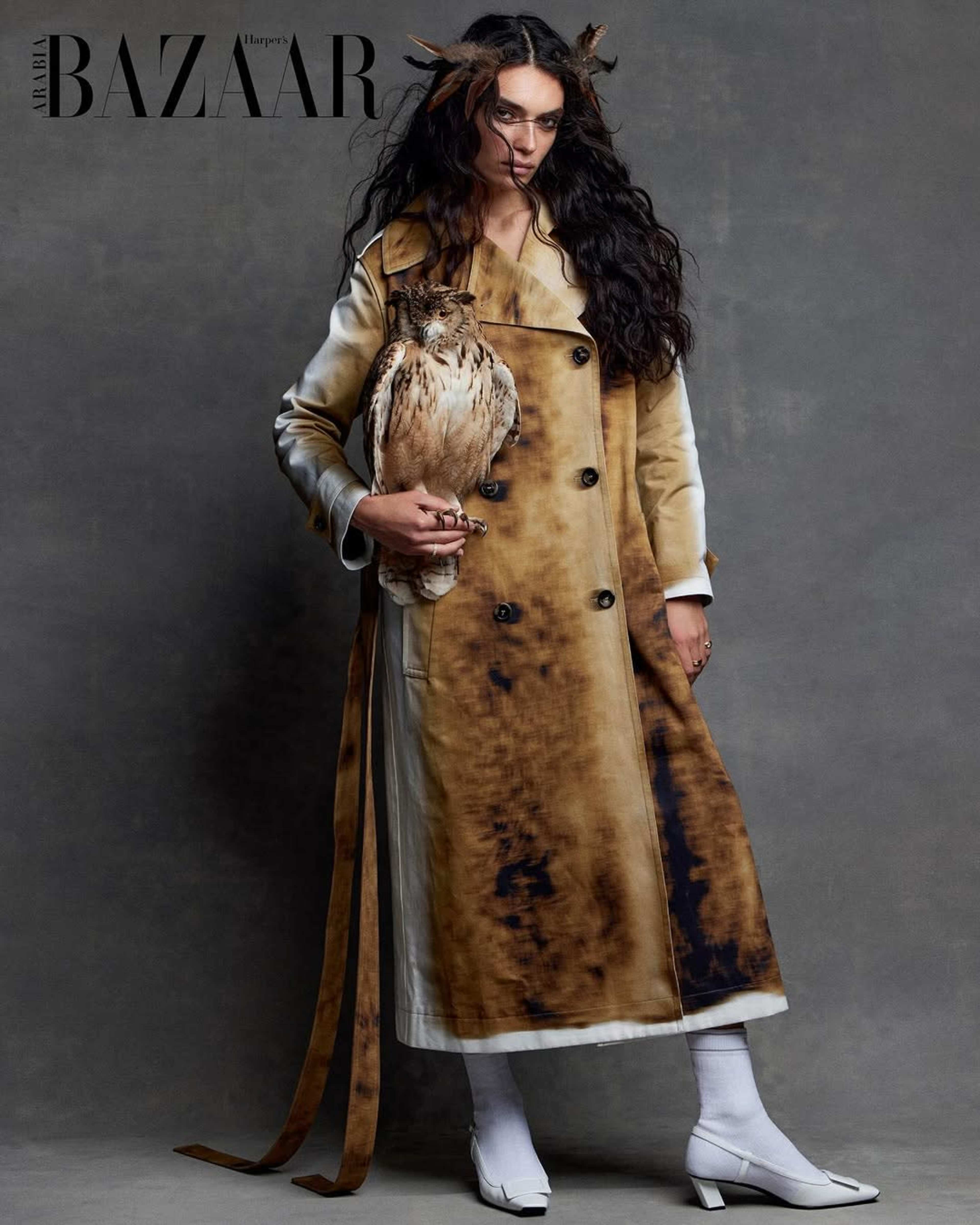 A model stands against a gray background wearing a long, two-tone coat while holding an owl.