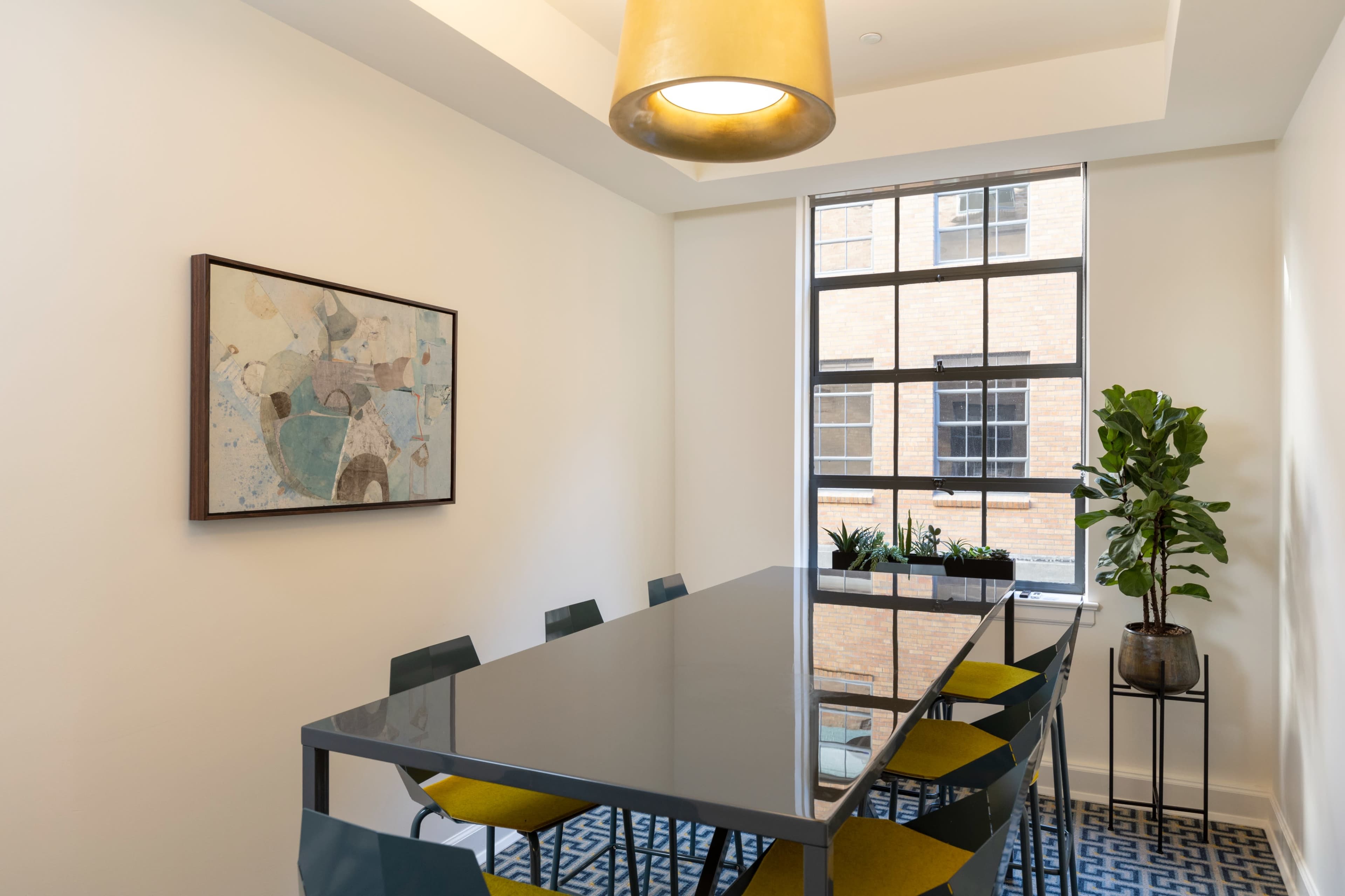 A modern conference room features a long glass table surrounded by colorful chairs, a large window with potted plants, and an abstract painting on the wall.