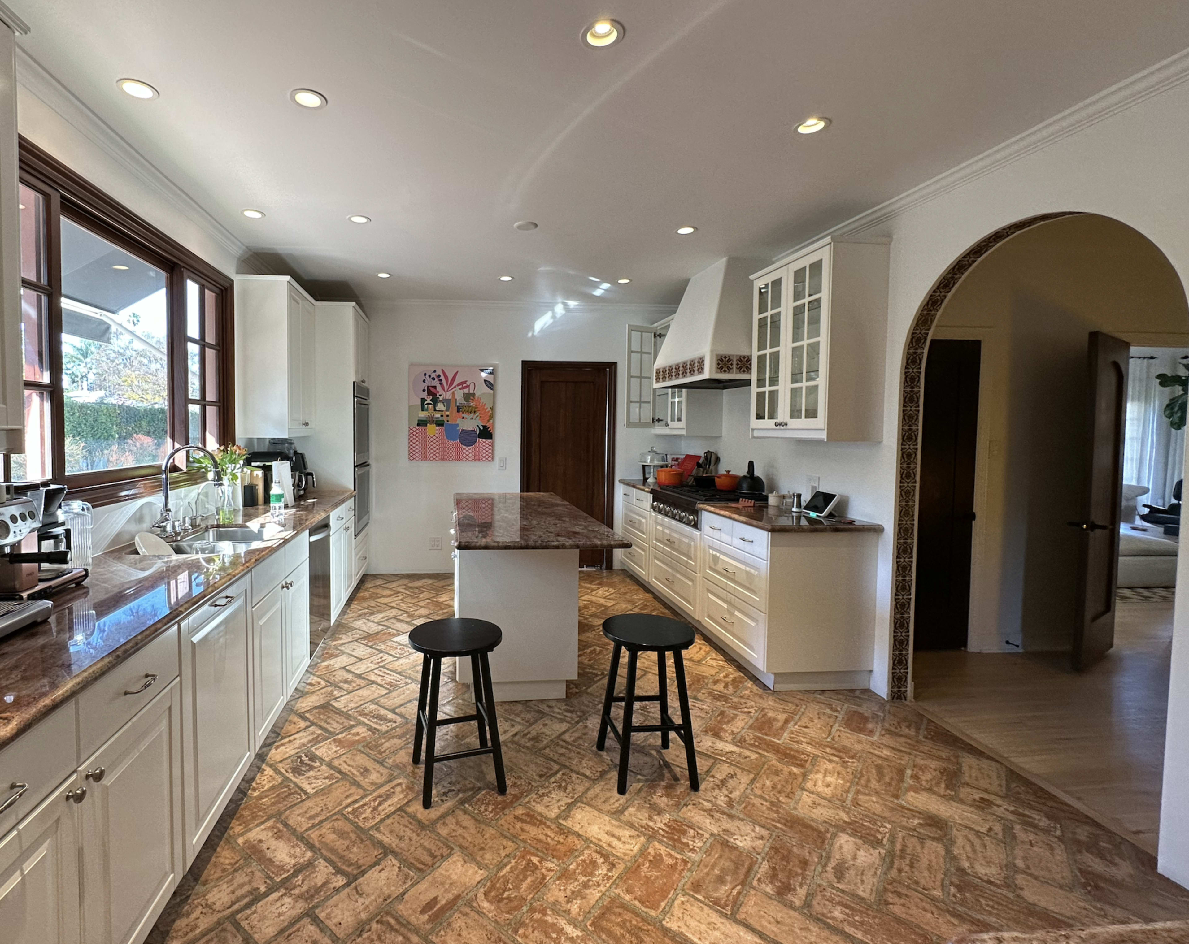 A spacious kitchen featuring white cabinetry, a large island with two black stools, and a tile floor with a herringbone pattern.