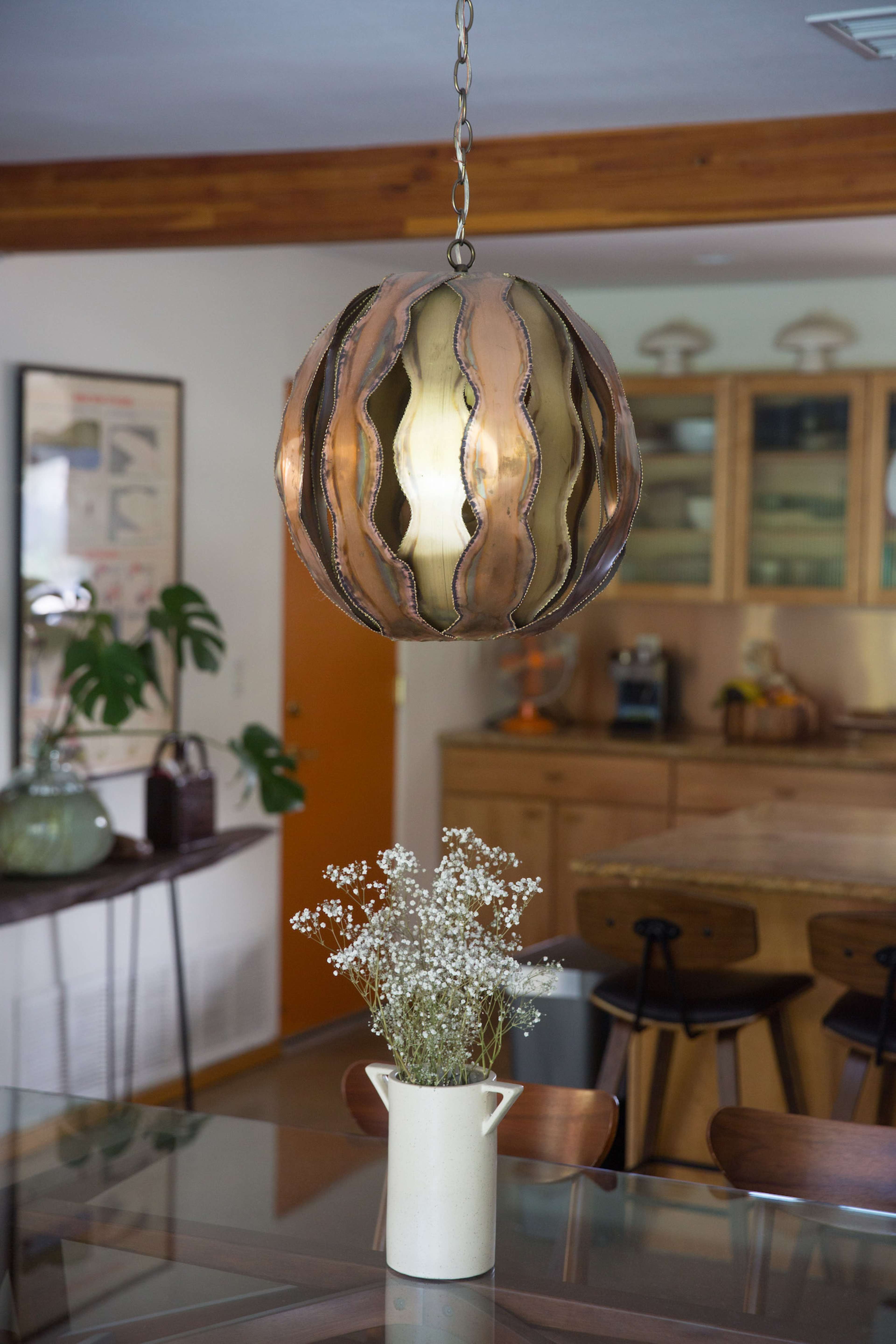 A metal pendant lamp hangs above a dining table with a small vase of flowers set on it, while an open kitchen area is visible in the background.