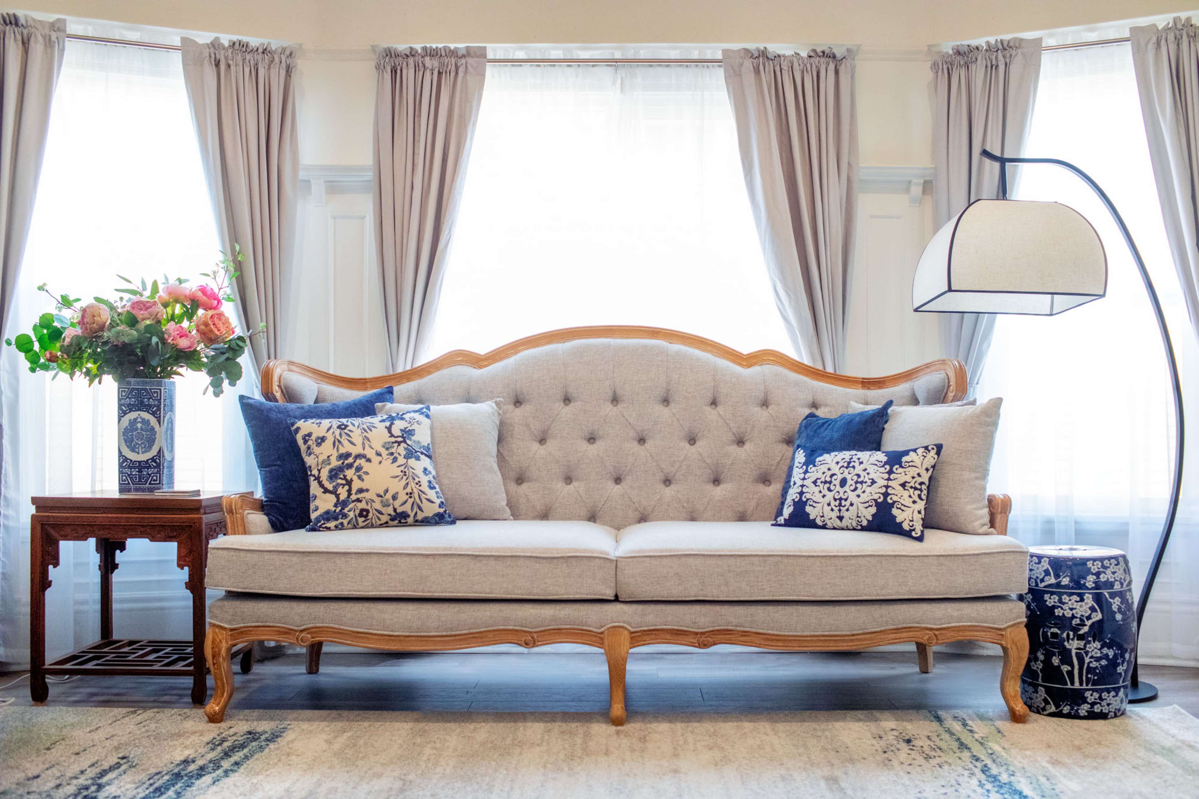 A gray tufted sofa with decorative pillows is positioned between two light-filtering windows, accompanied by a side table and a floor lamp.