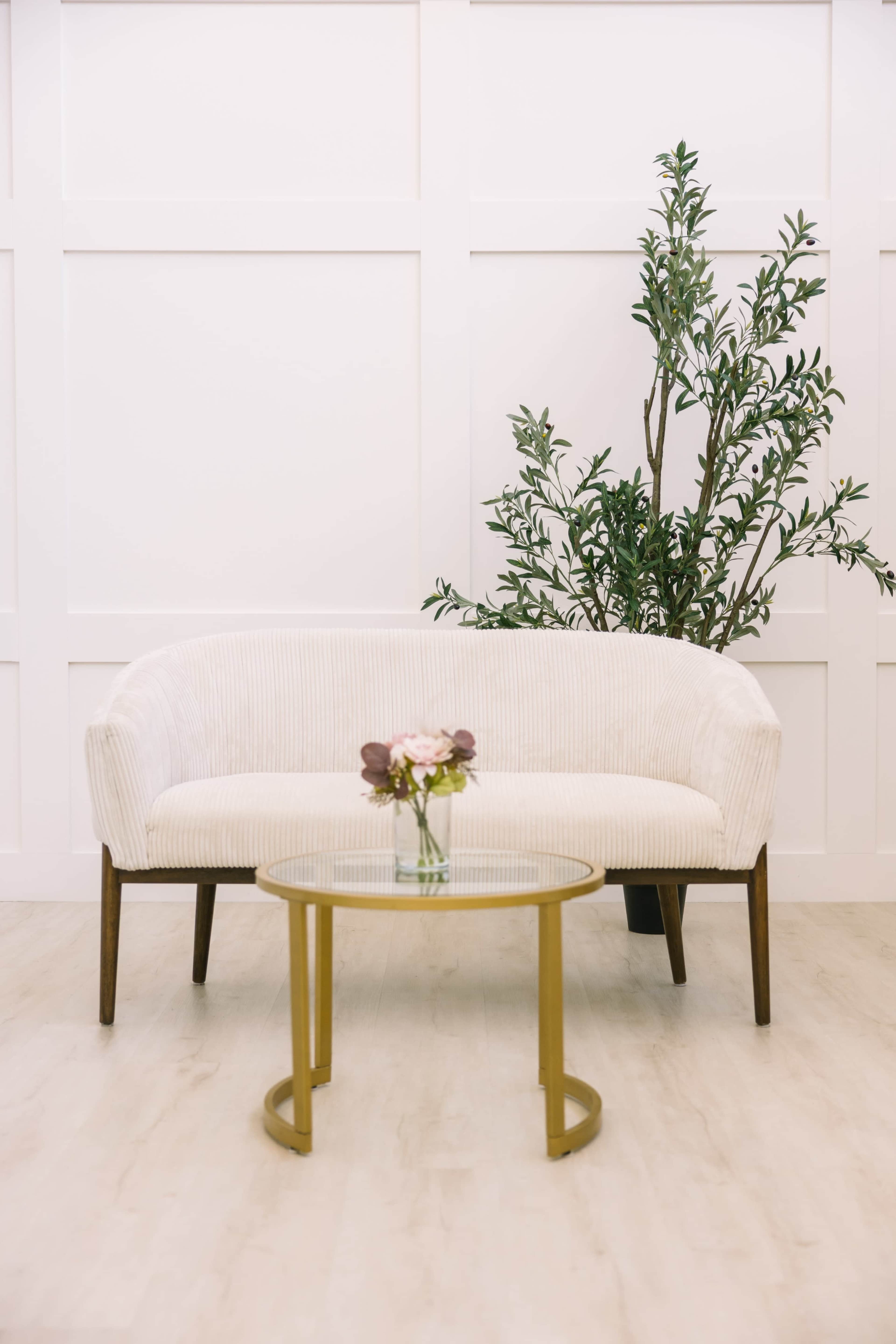 A white, curved sofa is positioned beside a small glass table adorned with a vase of flowers, contrasted against a minimalist backdrop with a potted plant.