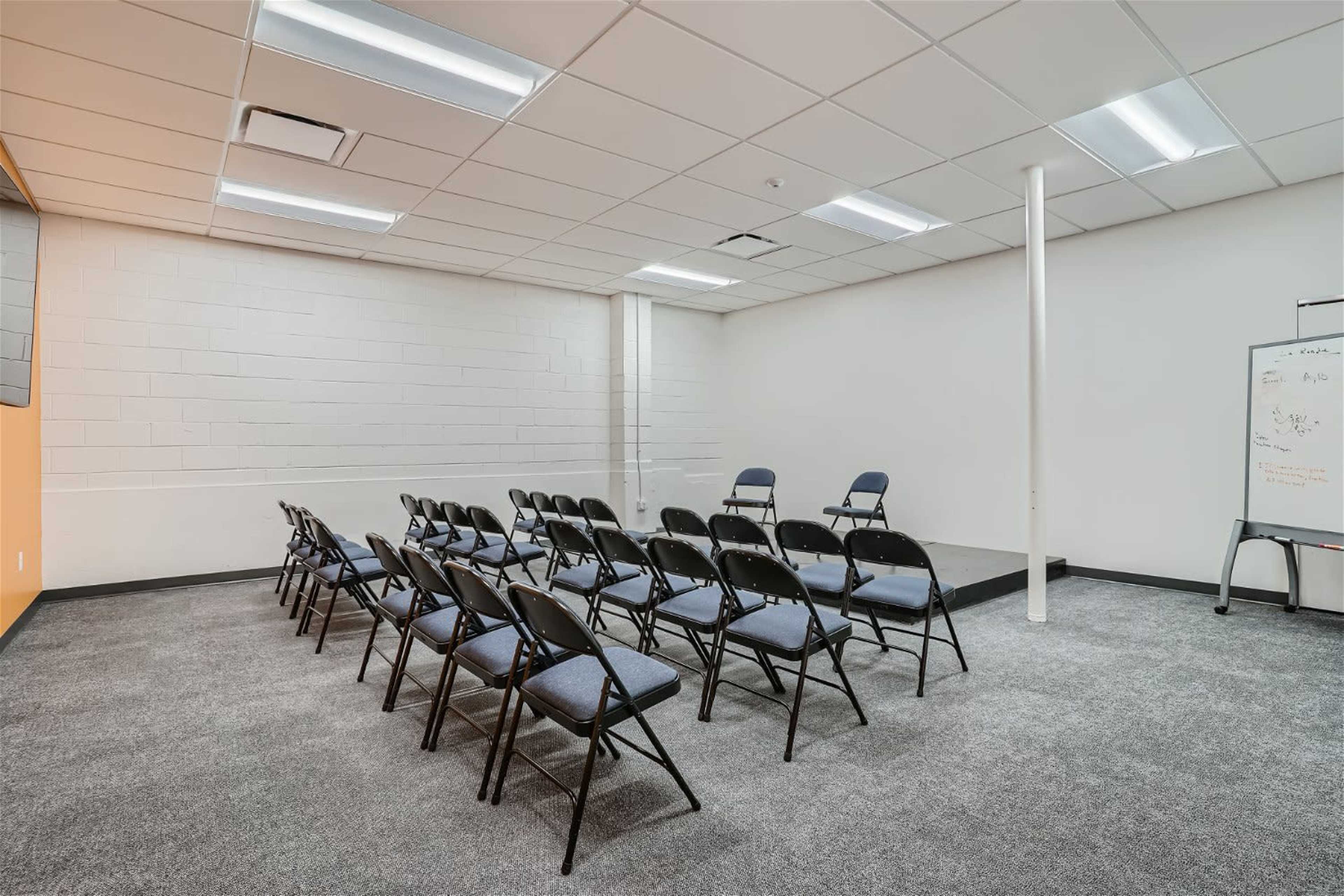 A small, empty meeting room features a row of black folding chairs facing a raised platform and a whiteboard against a wall.