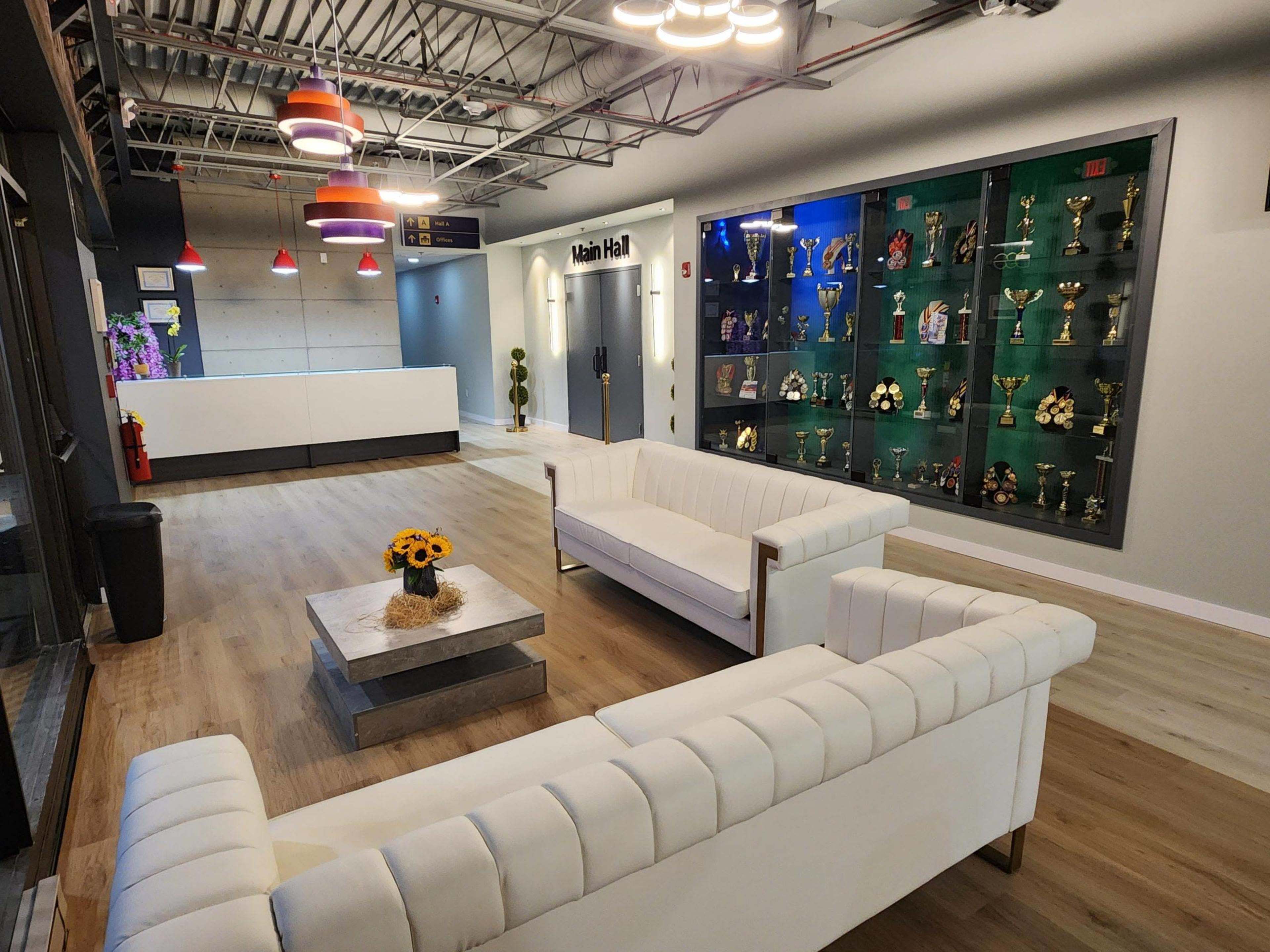 The image shows a modern lobby area featuring white sofas, a decorative coffee table with a flower arrangement, and a display wall filled with trophies and awards.