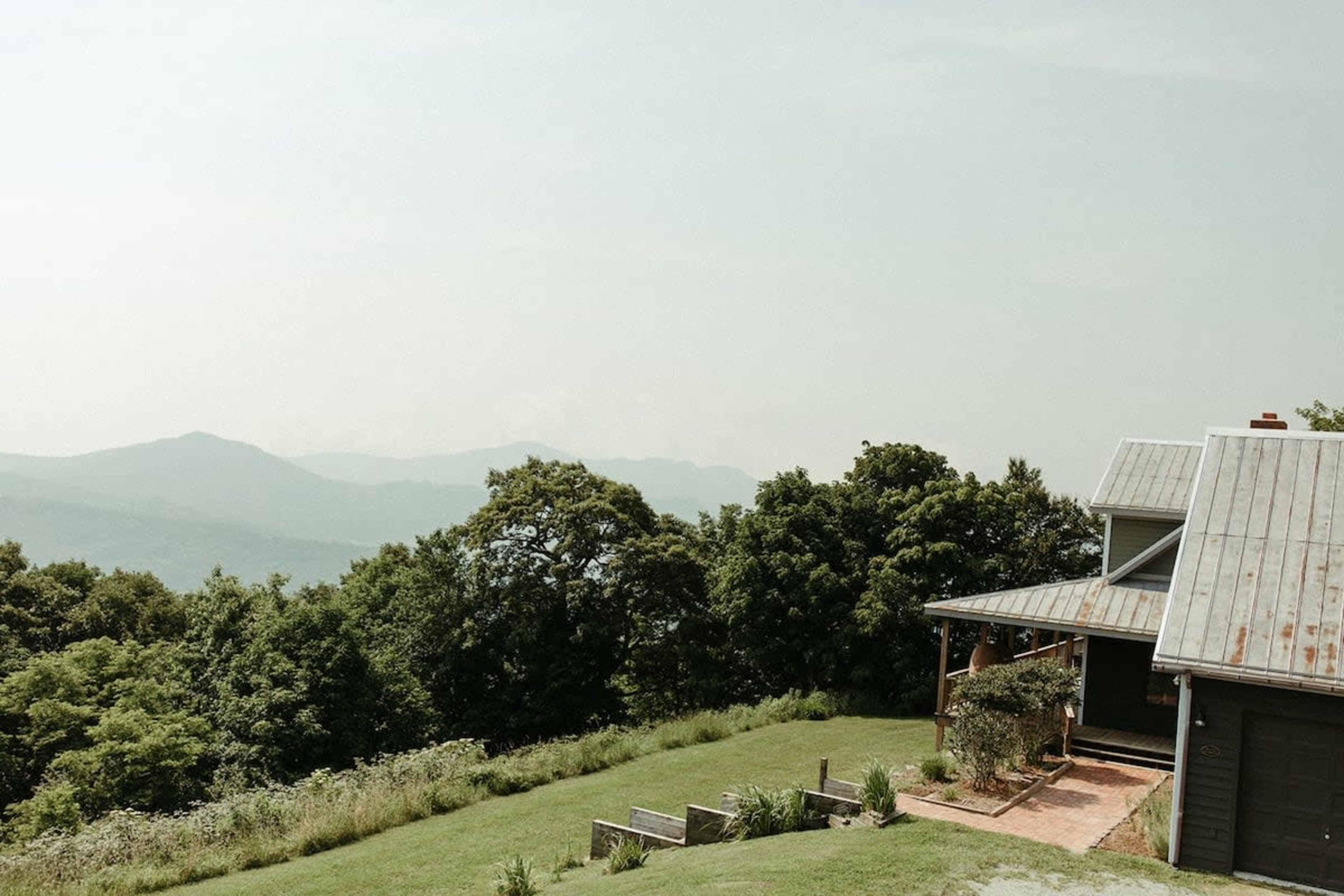 A house with a metal roof is situated on a hillside, overlooking a range of distant mountains and surrounded by trees and greenery.