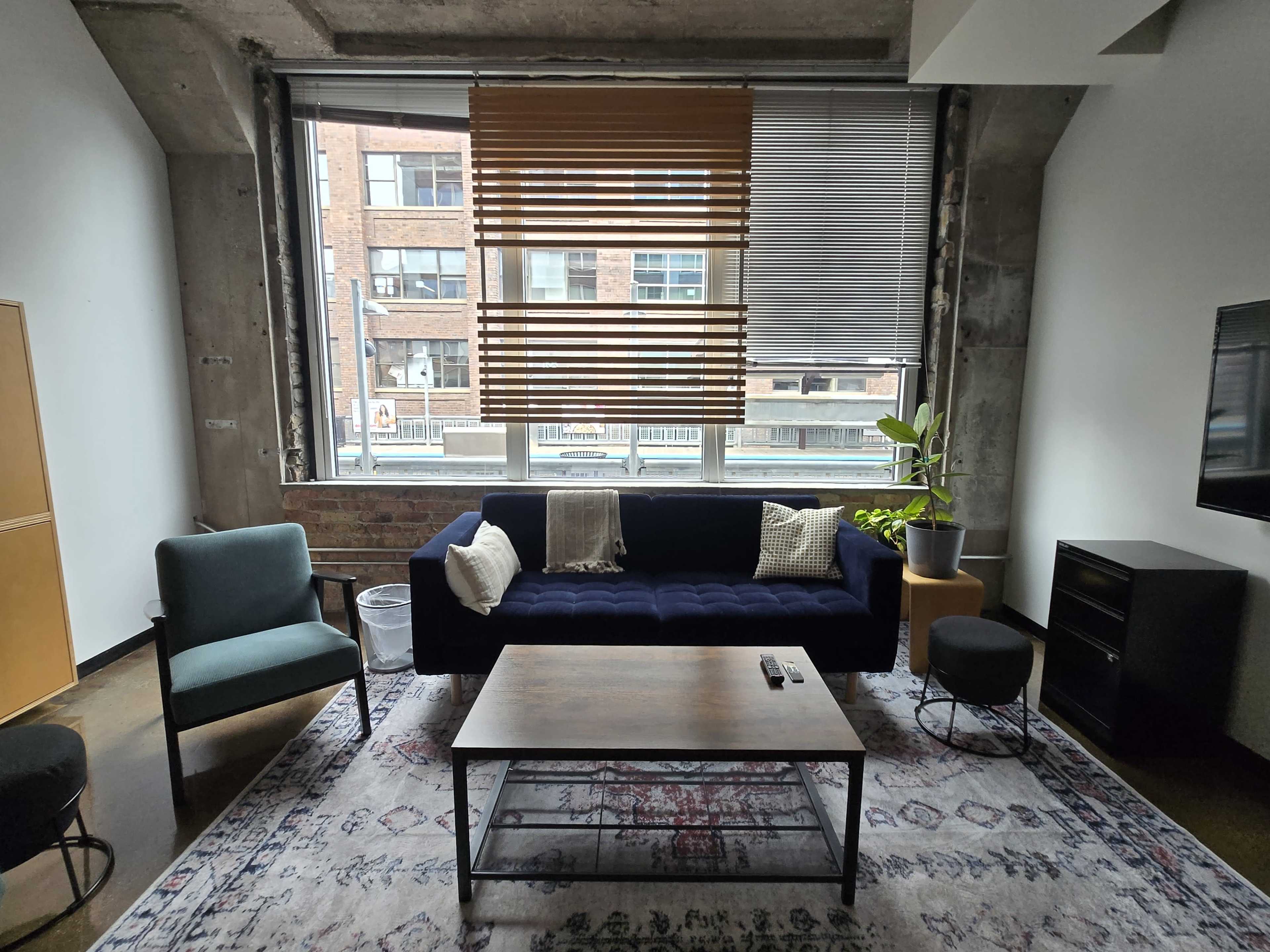 The image shows a modern living room with a blue sofa, a wooden coffee table, and large windows covered with blinds, featuring an indoor plant in the corner.