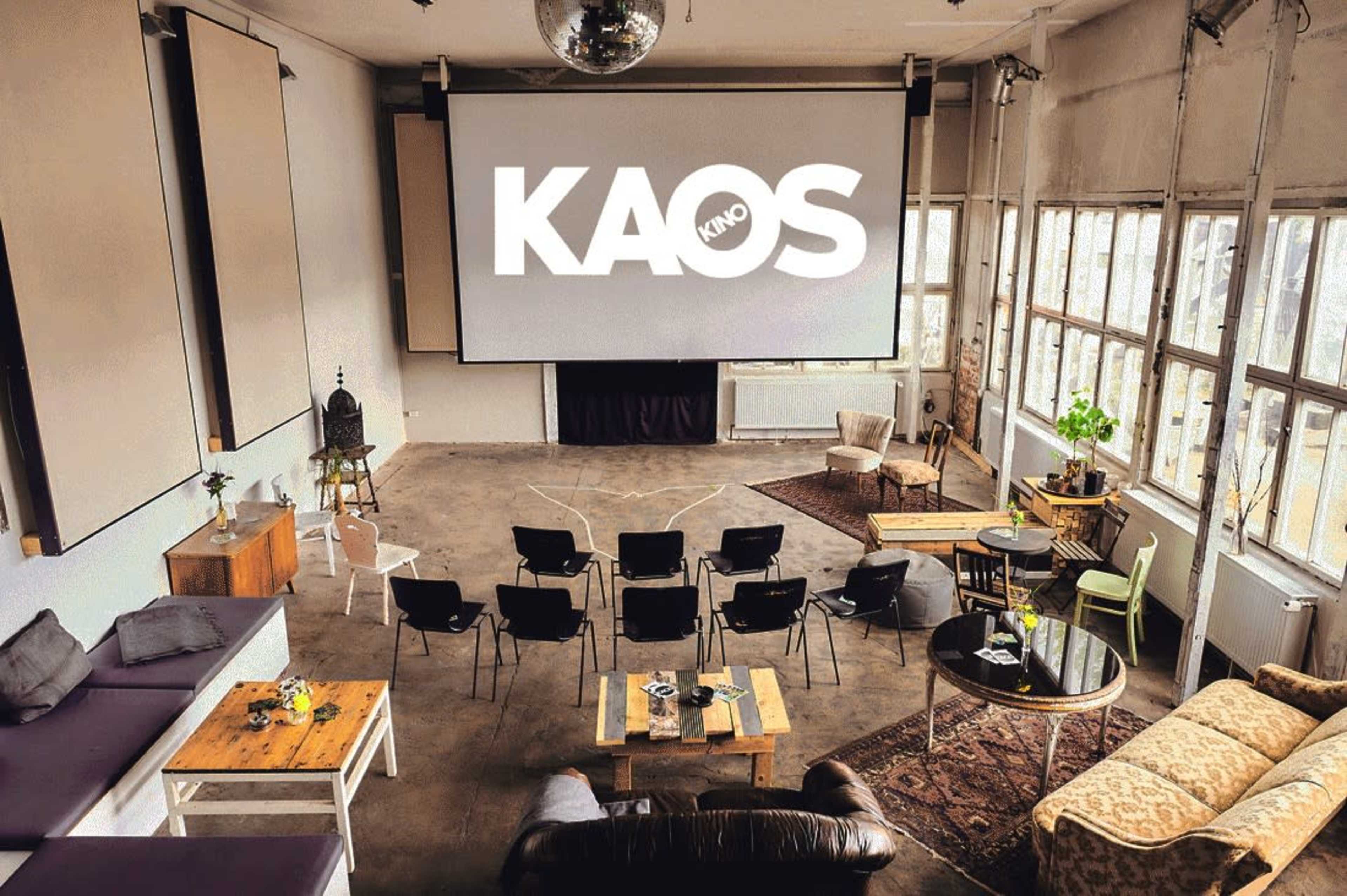 The image shows a spacious indoor venue with a large screen displaying "KAOS KINO," surrounded by various seating options including chairs and couches.