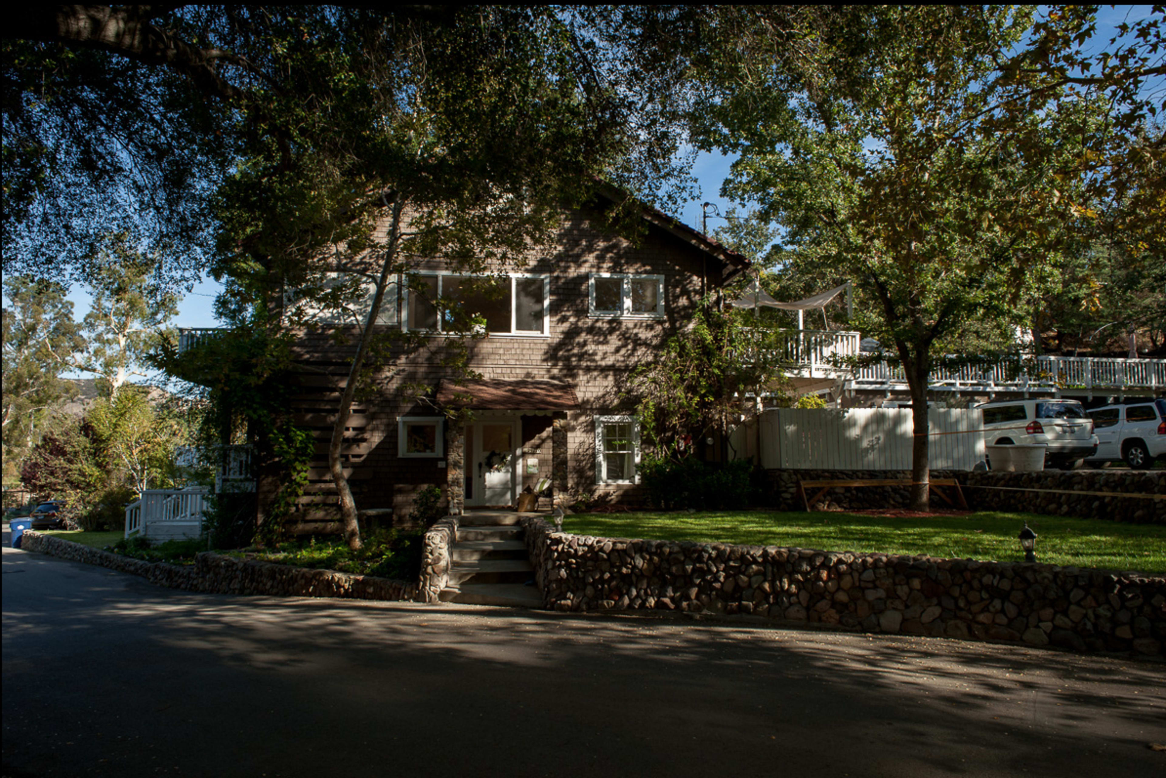 A two-story house with a stone wall is situated on a tree-lined street, surrounded by parked cars and greenery.