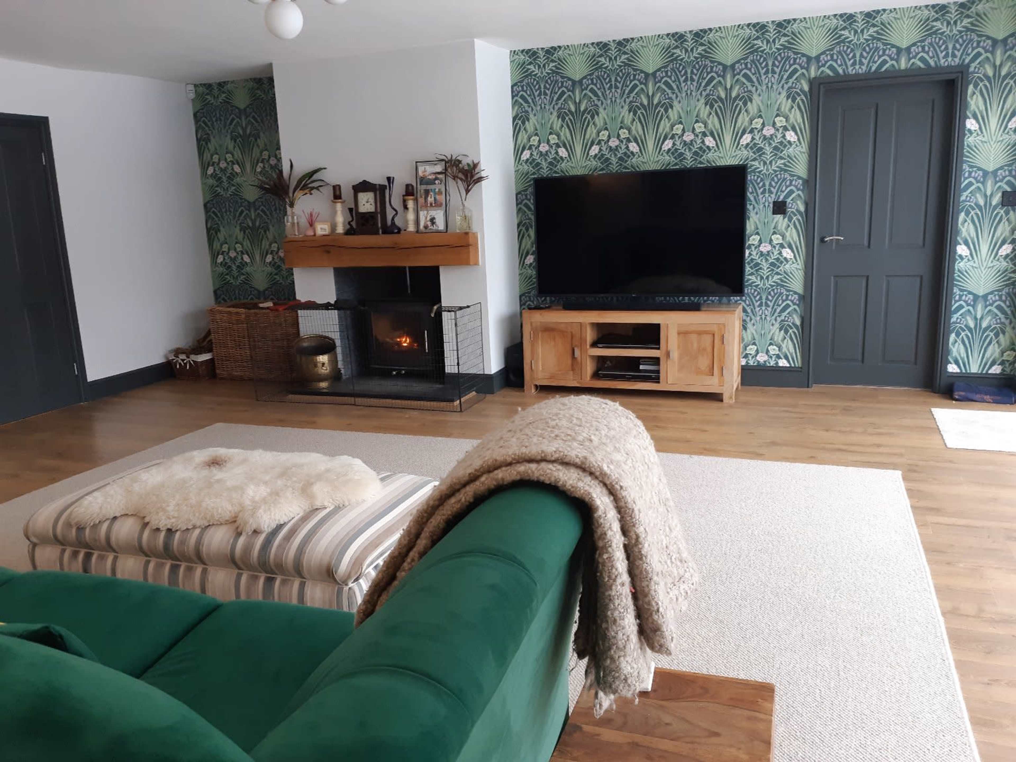 A living room featuring a green sofa, a wooden TV unit, a fireplace with a mantel, and patterned wallpaper with leaves and flowers.
