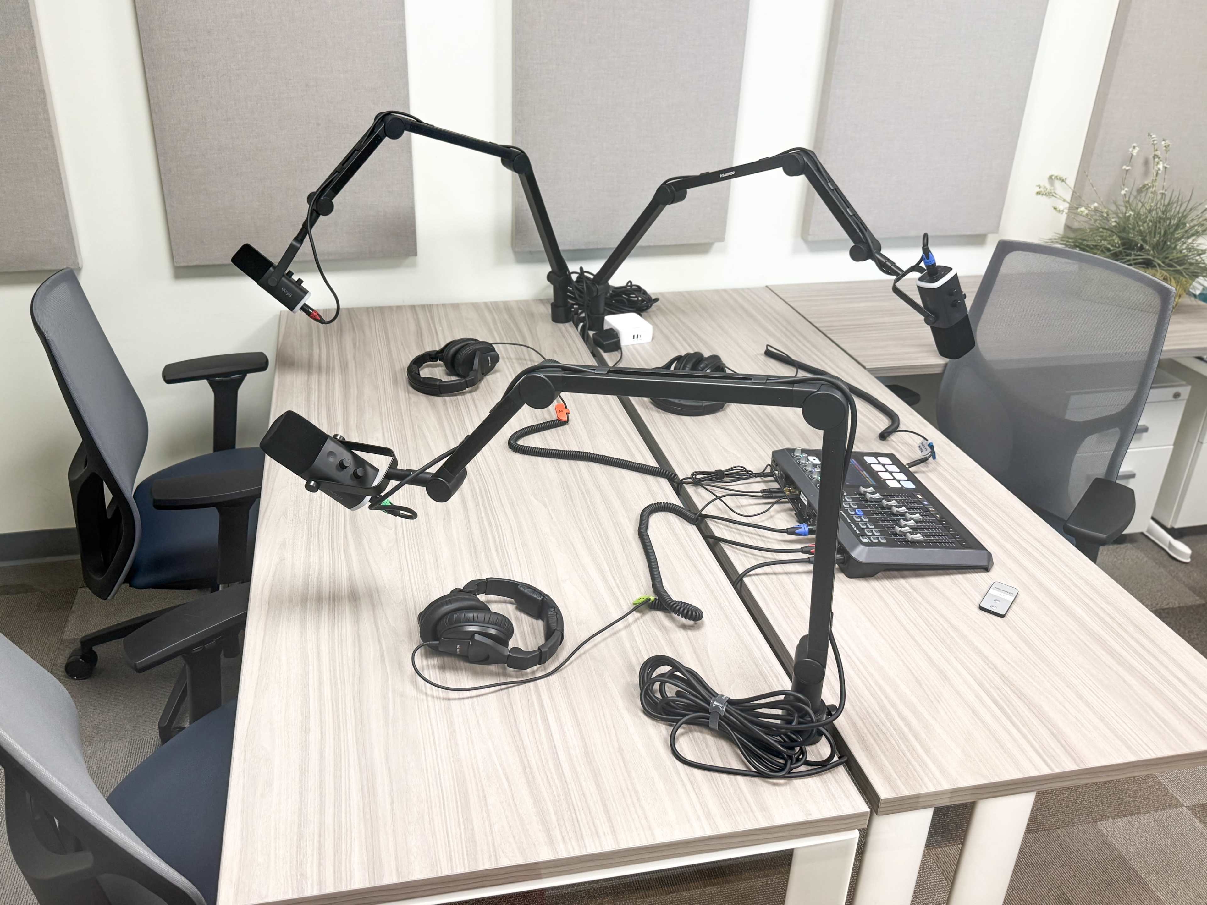 The image shows a recording studio setup with two adjustable microphone arms, a sound mixer, and headphones on a wooden table with chairs on either side.