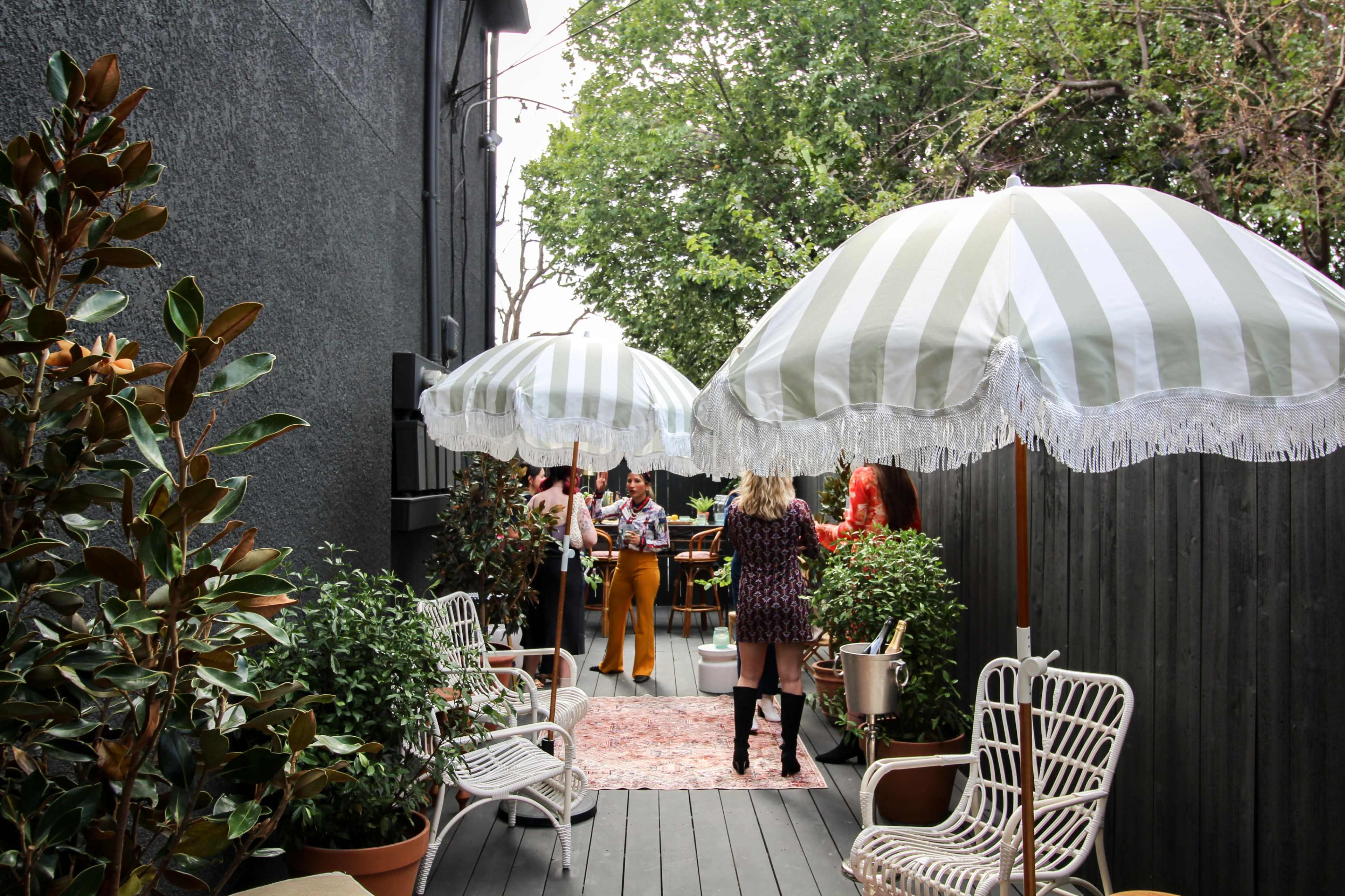 A narrow outdoor space lined with potted plants and patio furniture, featuring striped umbrellas overhead and people gathered in the background.