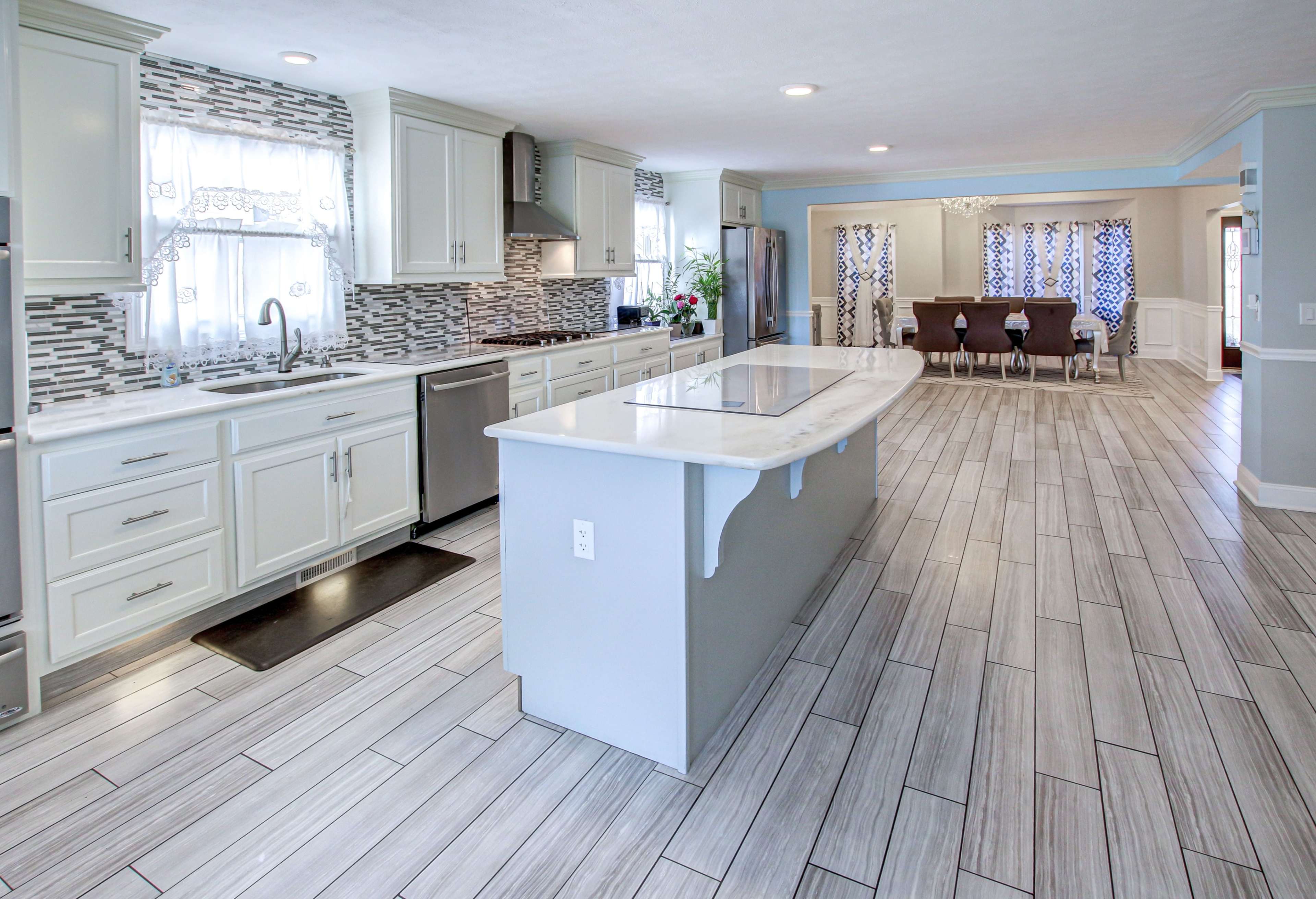 A modern kitchen with white cabinetry, a large island, and tiled flooring, leading to a dining area with a table and chairs.
