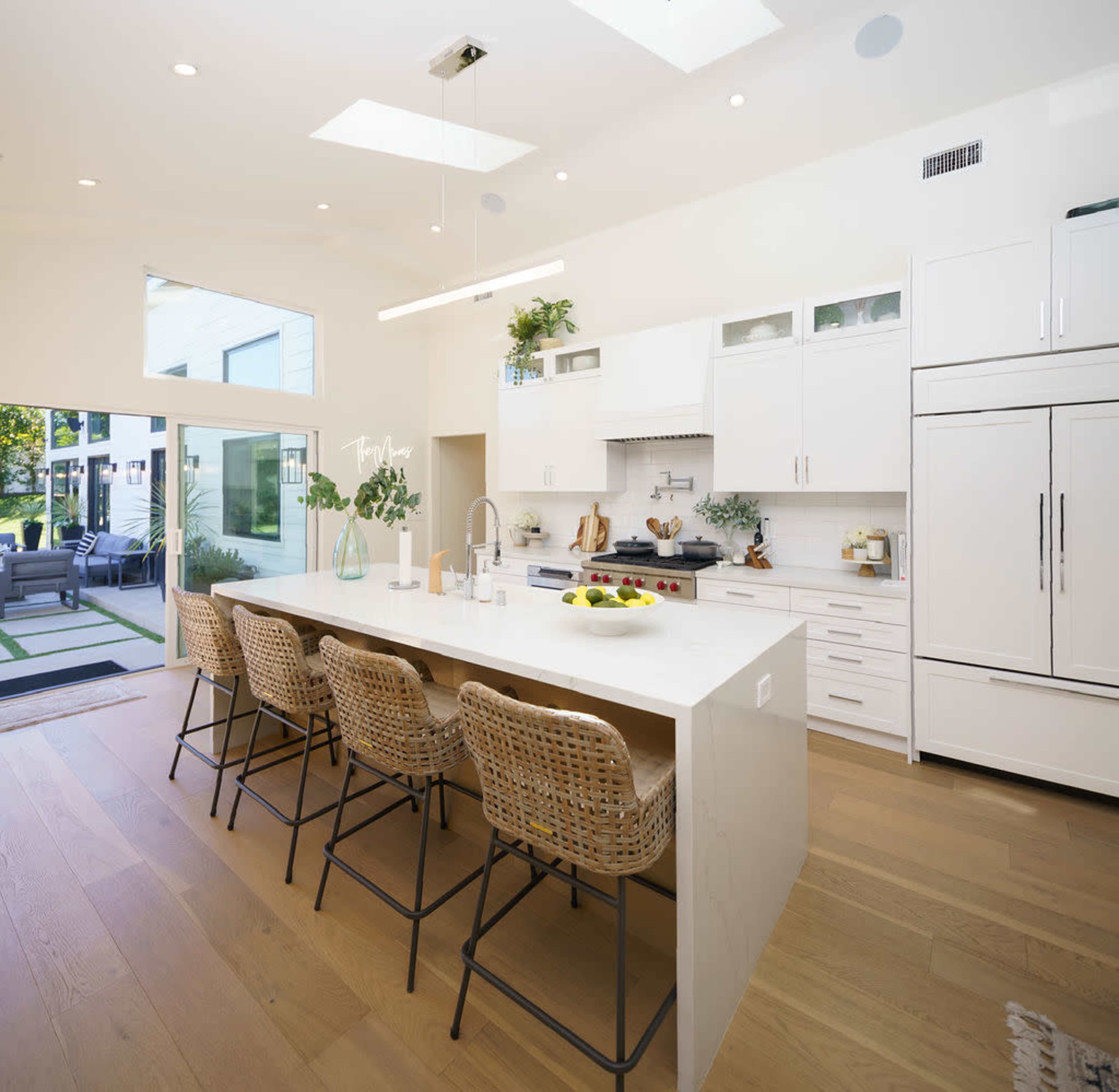 A modern kitchen features a large island with bar seating, white cabinetry, a gas stove, and large windows providing natural light.