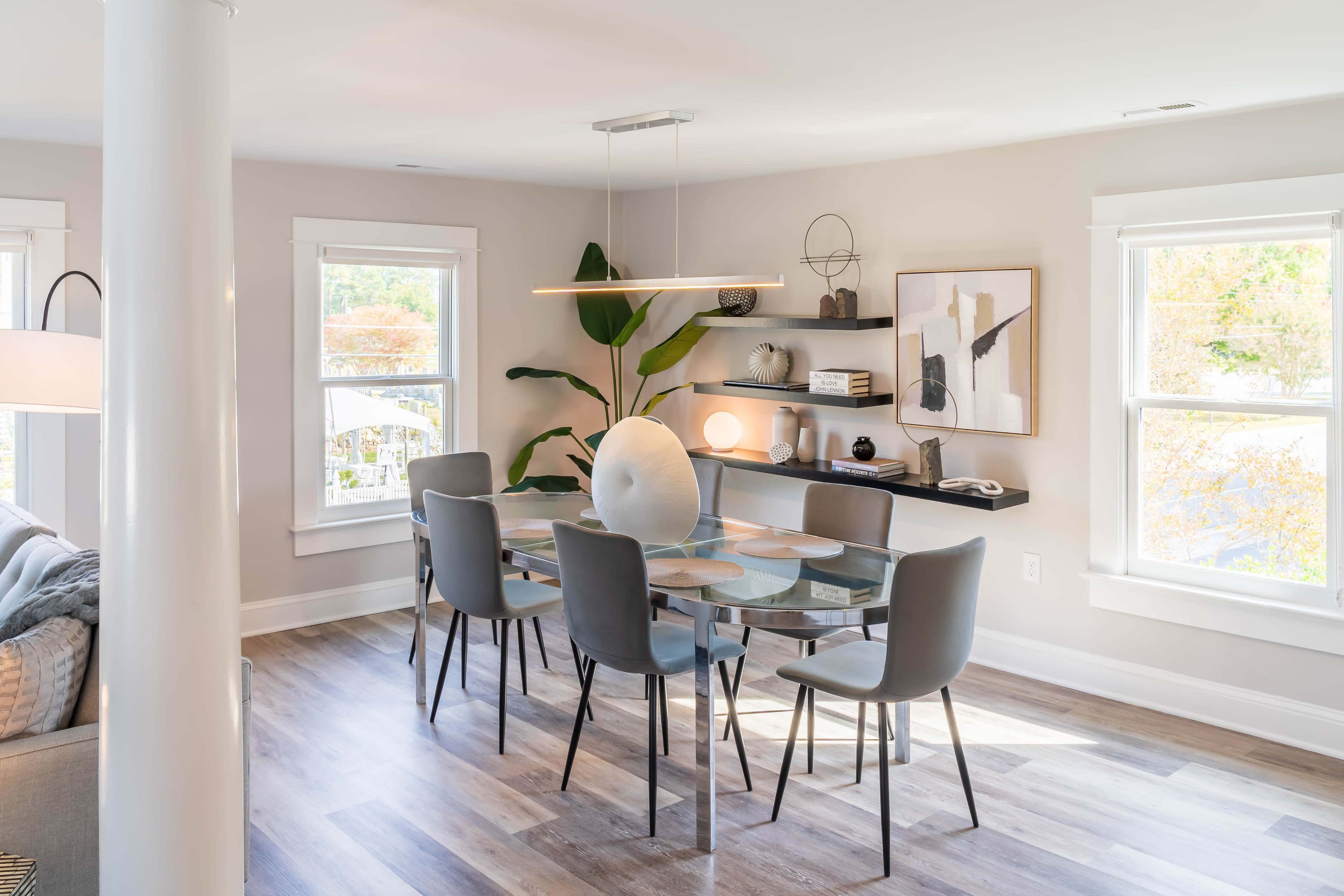 A modern dining area features a glass table surrounded by gray chairs, with a pendant light above and shelves displaying decorative items against a light-colored wall.