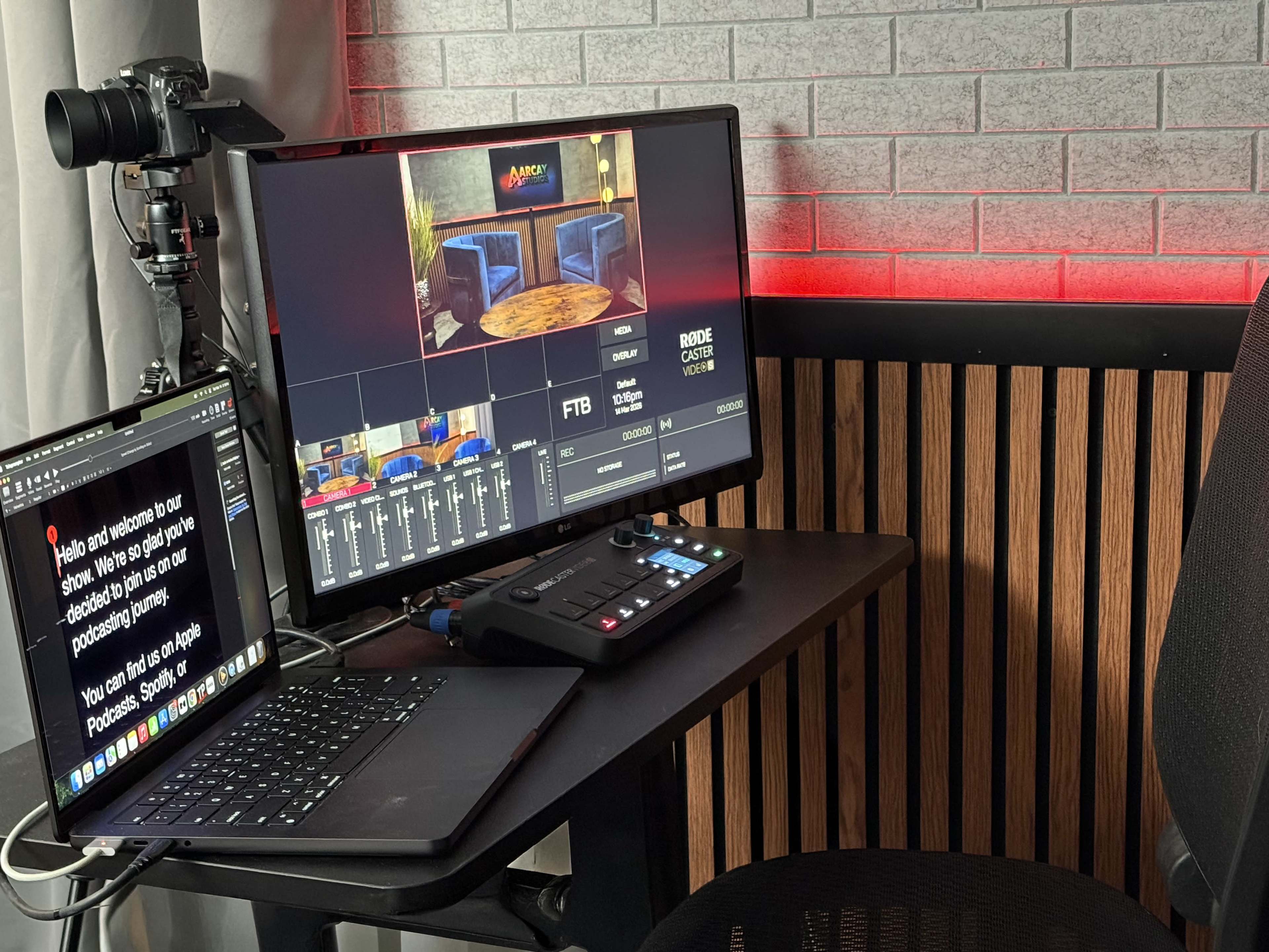 The image shows a neatly arranged desk setup with a laptop, a video production switcher, and a monitor displaying a control interface, all in a well-lit room with red accent lighting.