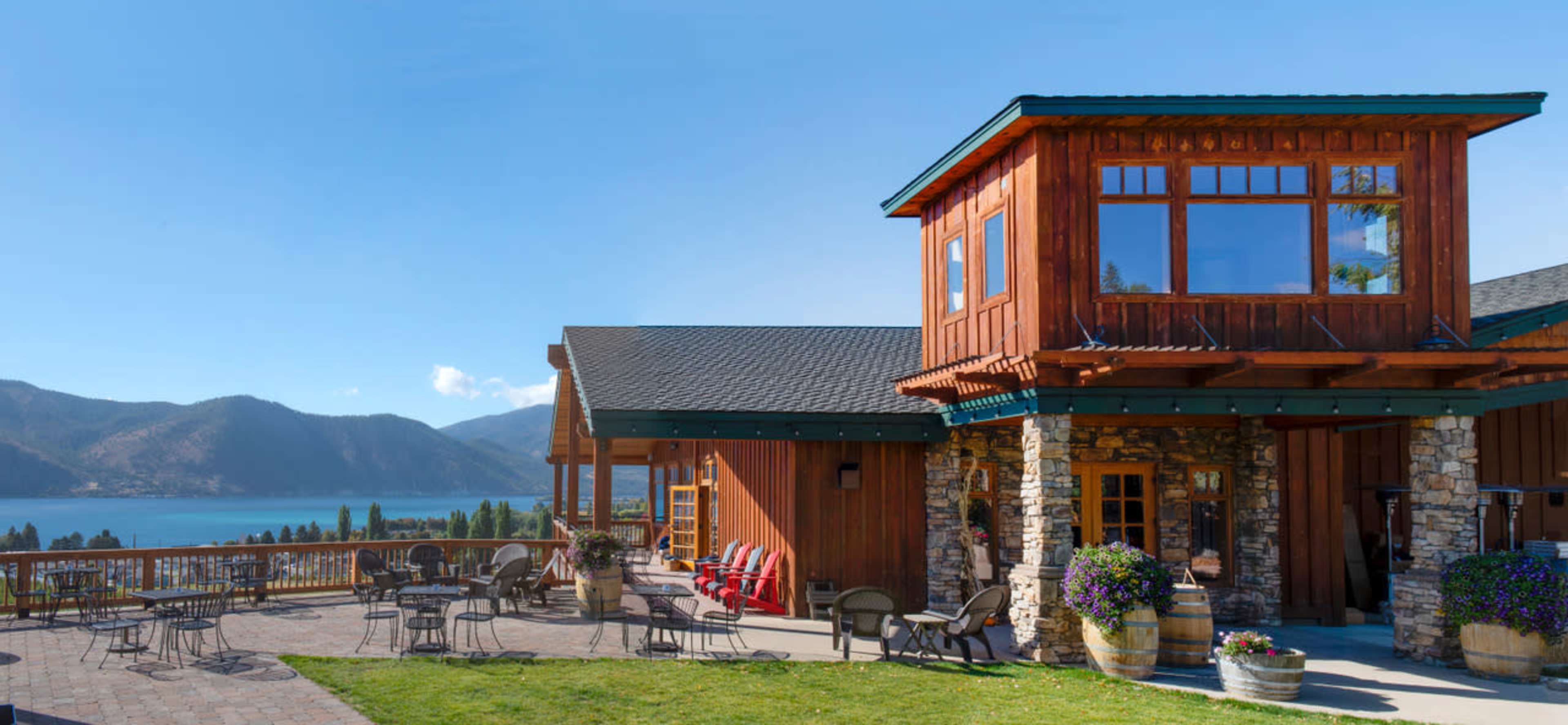 A wooden lodge with large windows overlooks a lake surrounded by mountains, with patio seating in the foreground.