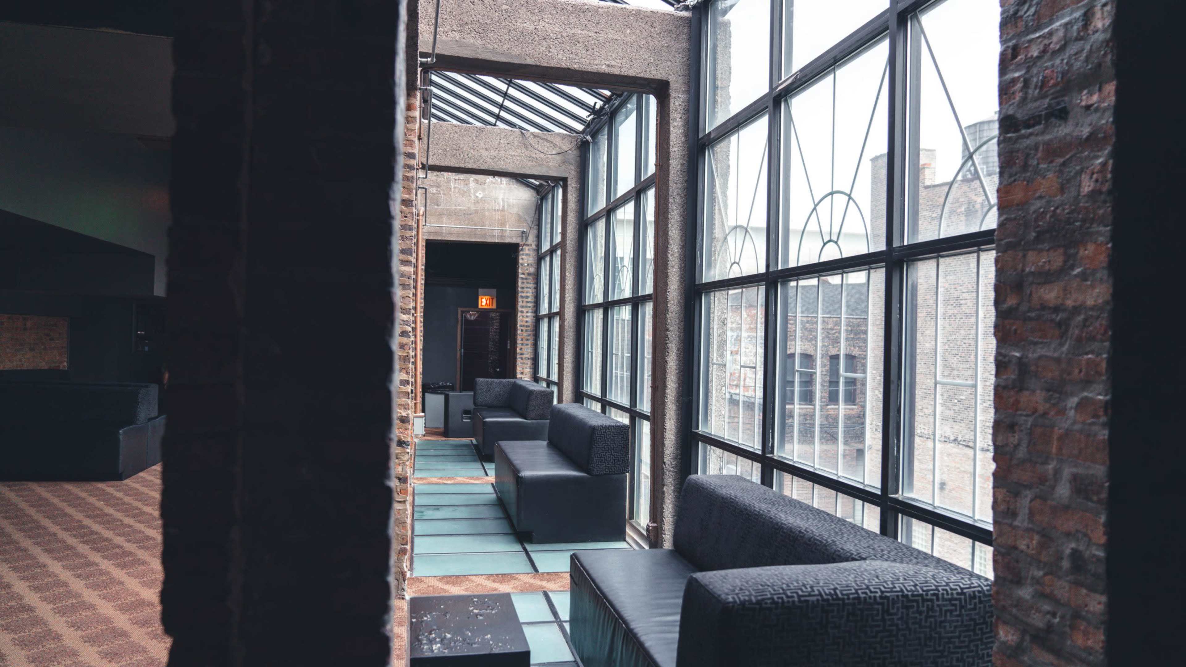 The image shows a modern lounge area with large windows, brick walls, and black sofas arranged along a narrow corridor.