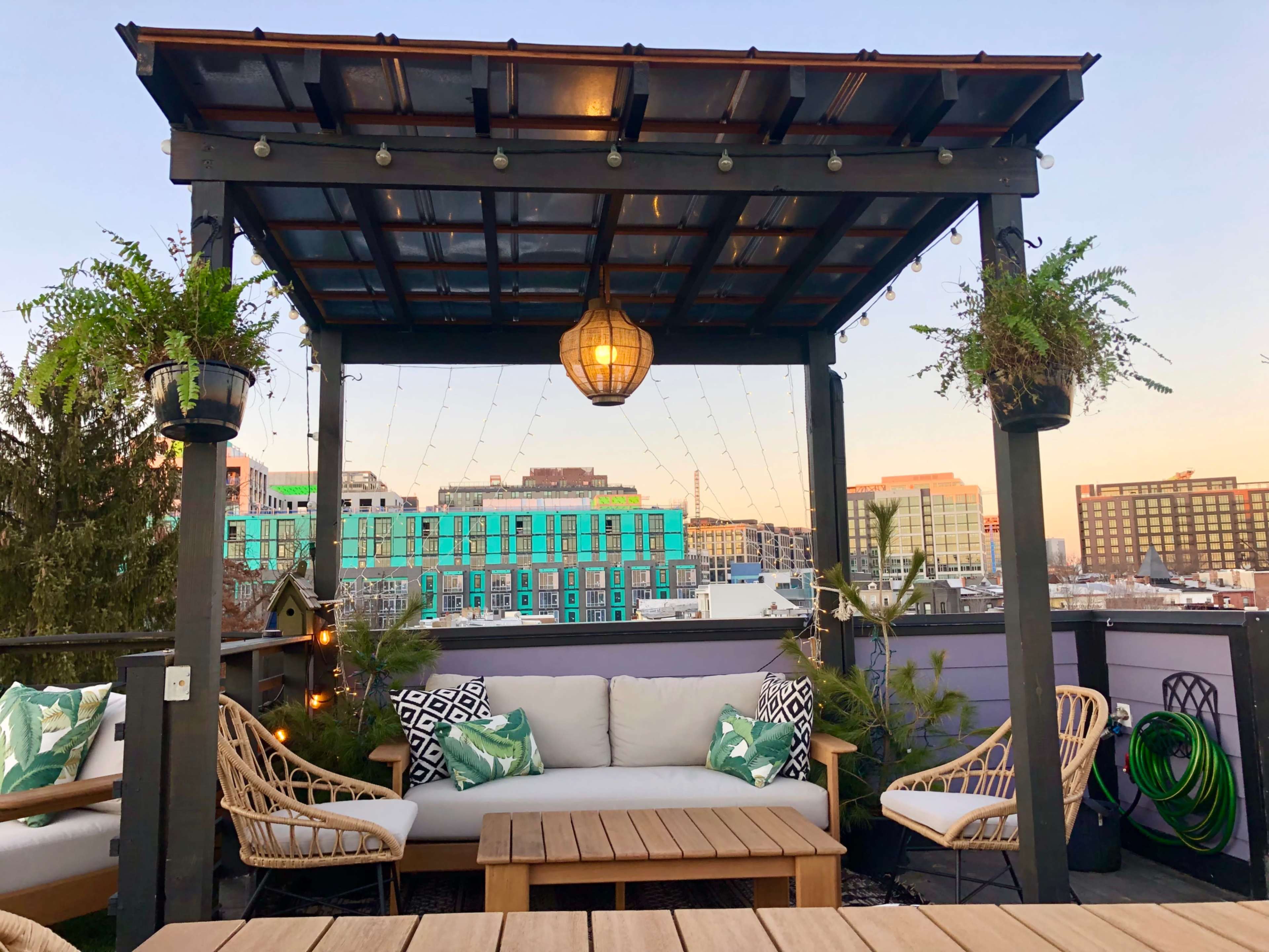 A rooftop deck features a seating area under a wooden pergola adorned with hanging plants and string lights, overlooking a city skyline.