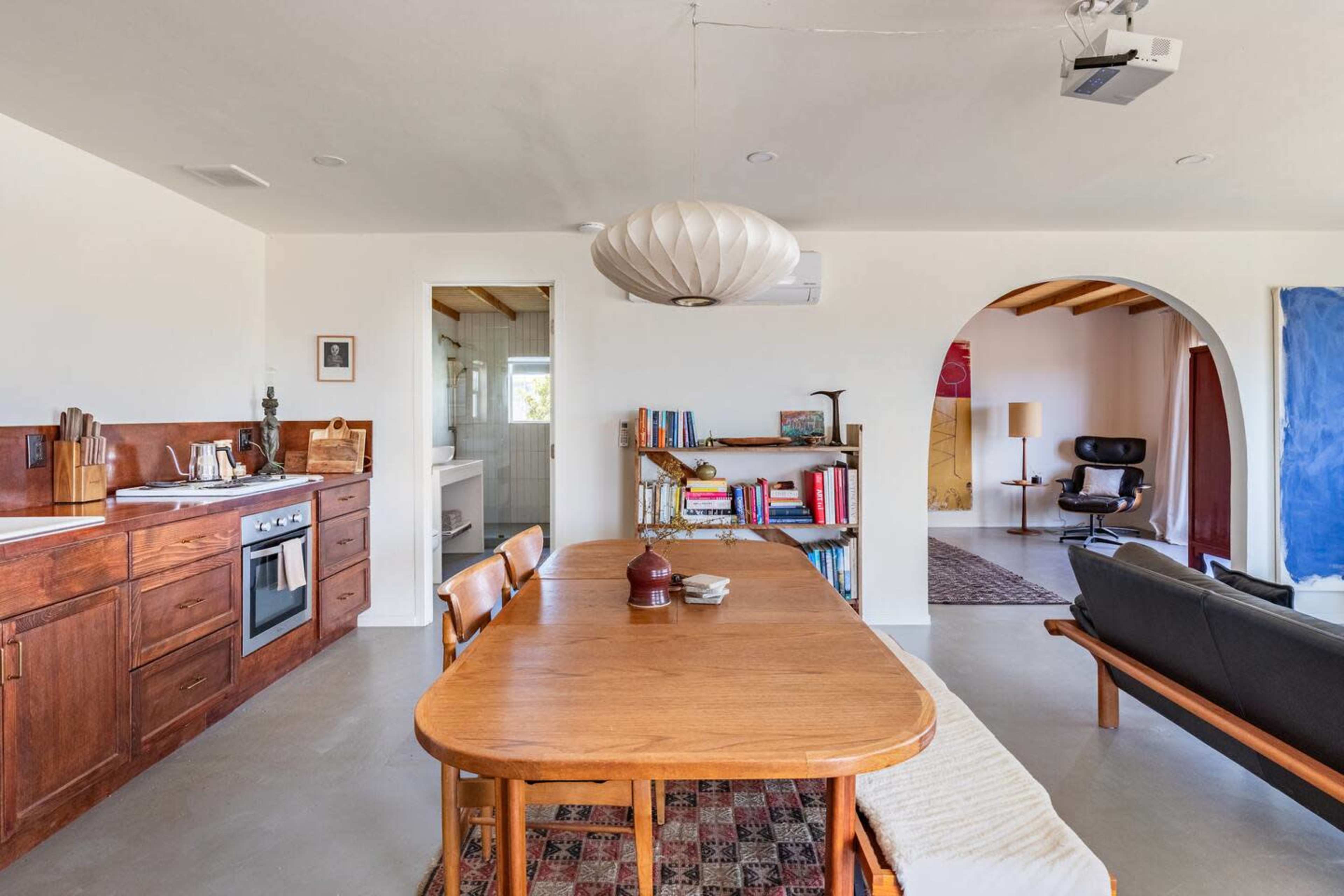 A modern kitchen and dining area features a wooden table, chairs, an oven, and shelves with books, adjacent to a living space with a sofa and a projection screen.