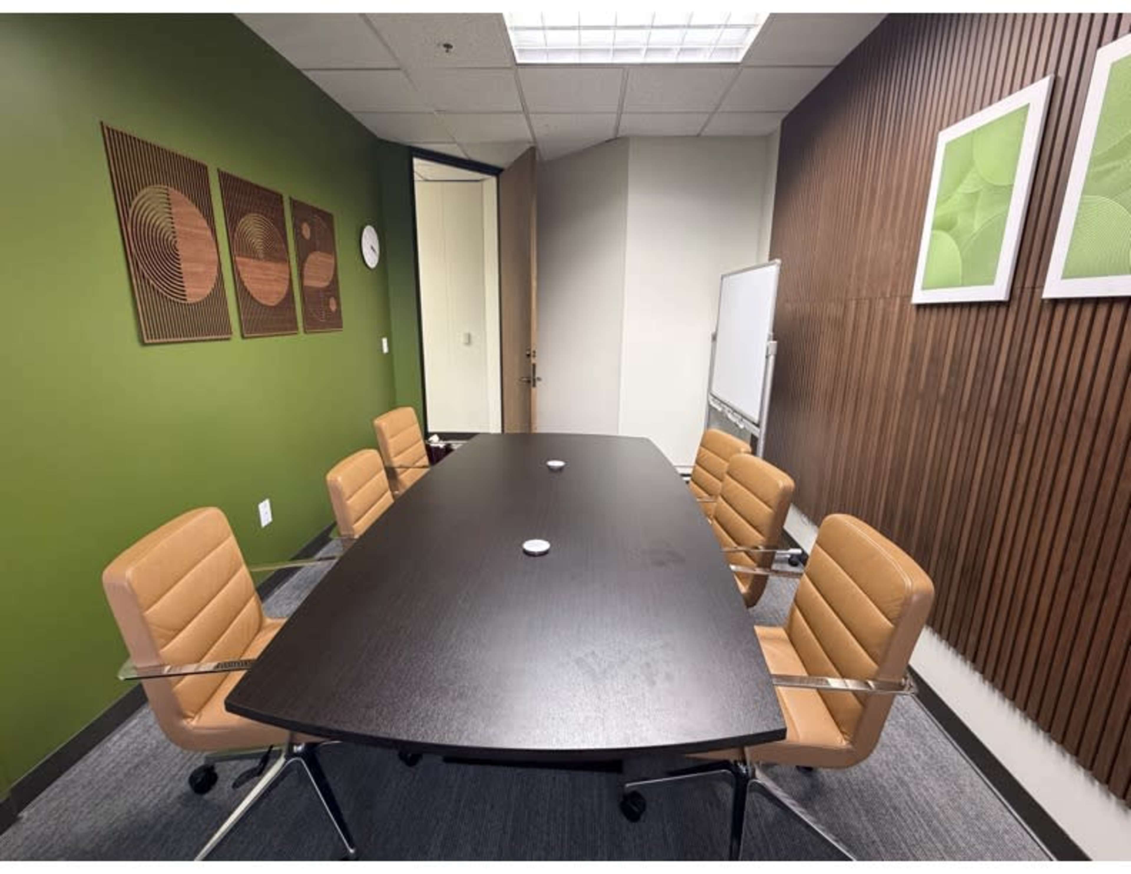 The image shows a small conference room with a dark rectangular table surrounded by four tan chairs, green walls, and modern artwork on the walls.