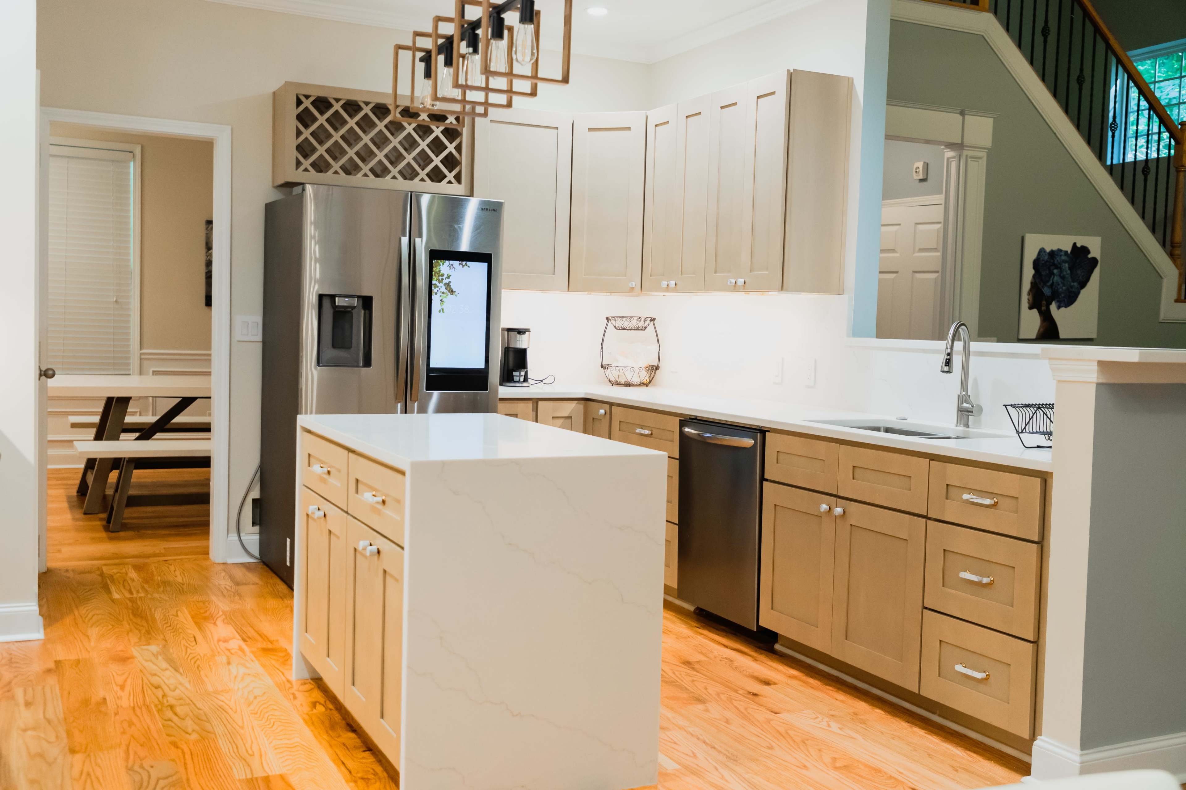 A modern kitchen with light wood cabinets, stainless steel appliances, a central island with a white countertop, and hardwood flooring.