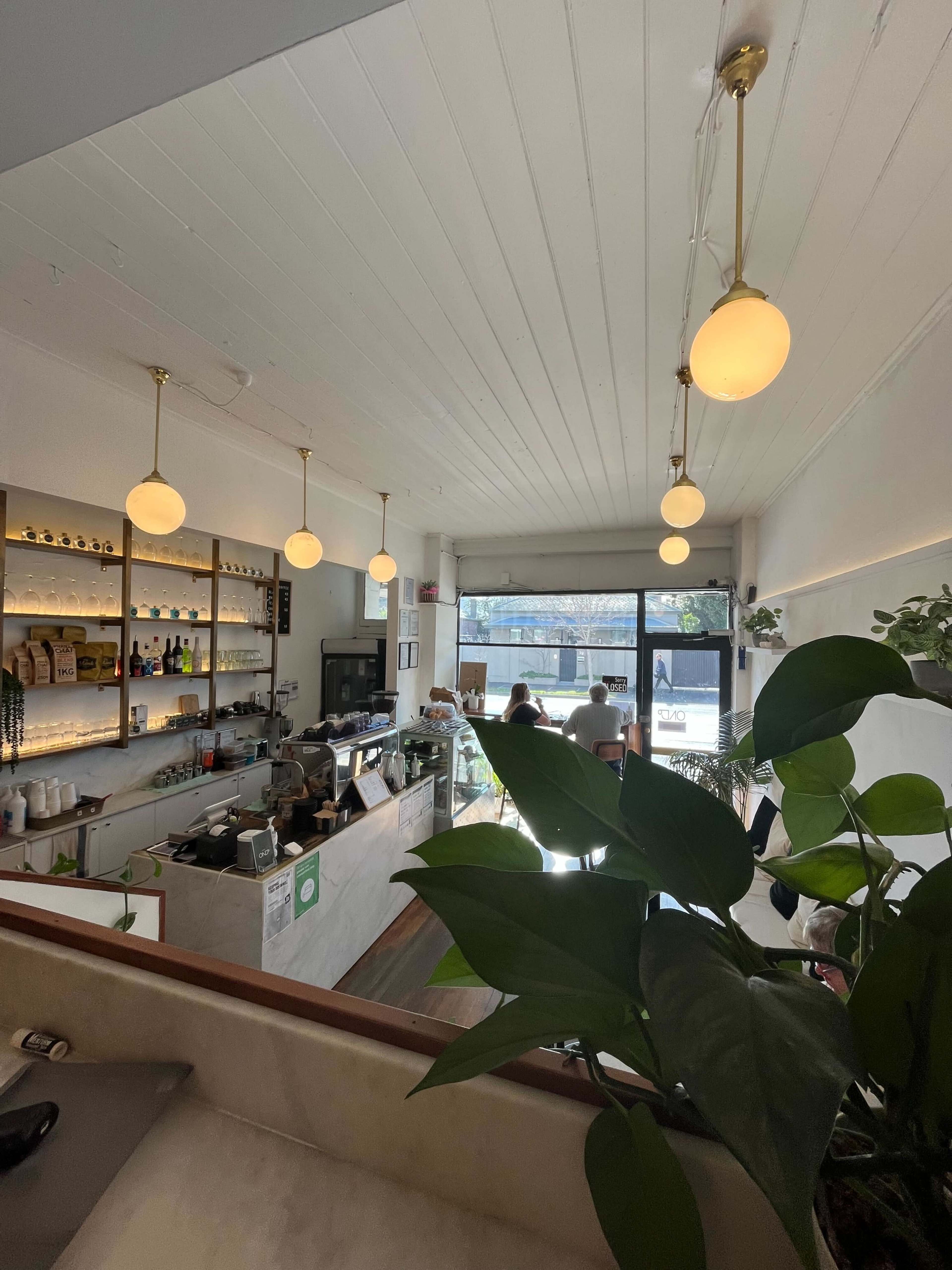 A café interior features a counter with shelves of beverages, pendant lights, and large windows allowing natural light to illuminate the space.