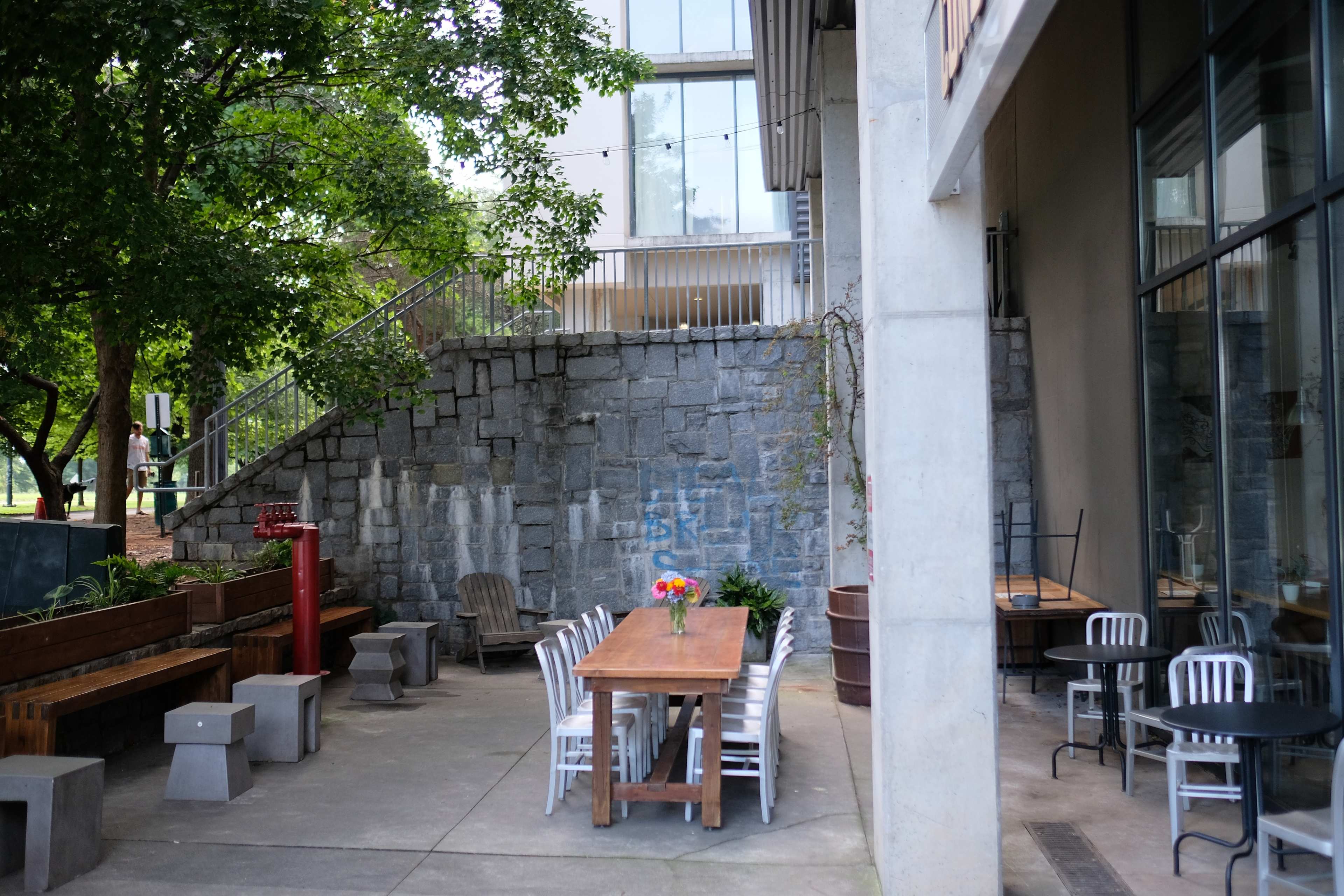 An outdoor dining area features a long wooden table with flowers surrounded by benches and metal chairs against a stone wall.