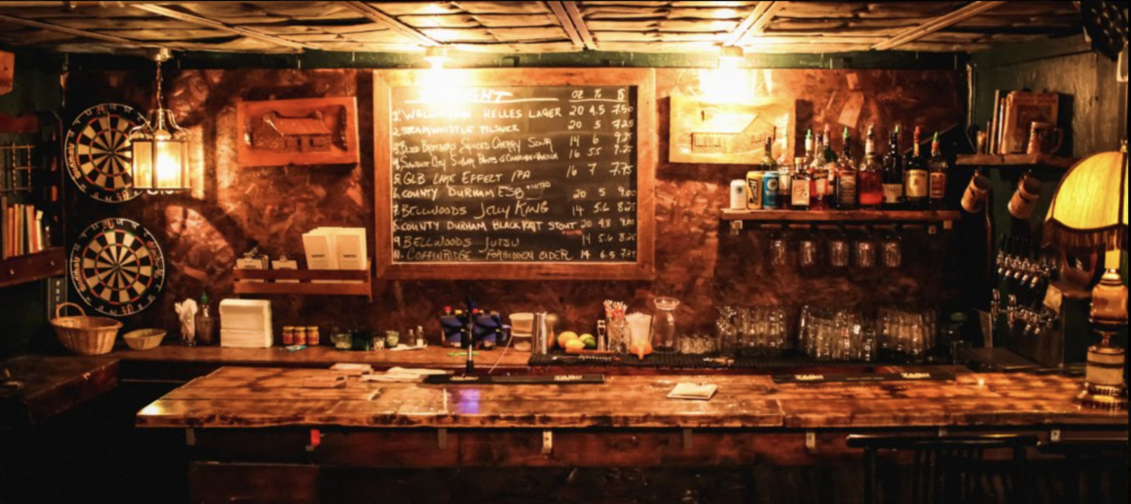 A wooden bar is set against a dark wall adorned with a chalkboard menu and shelves of various liquor bottles, while dartboards are visible on either side.