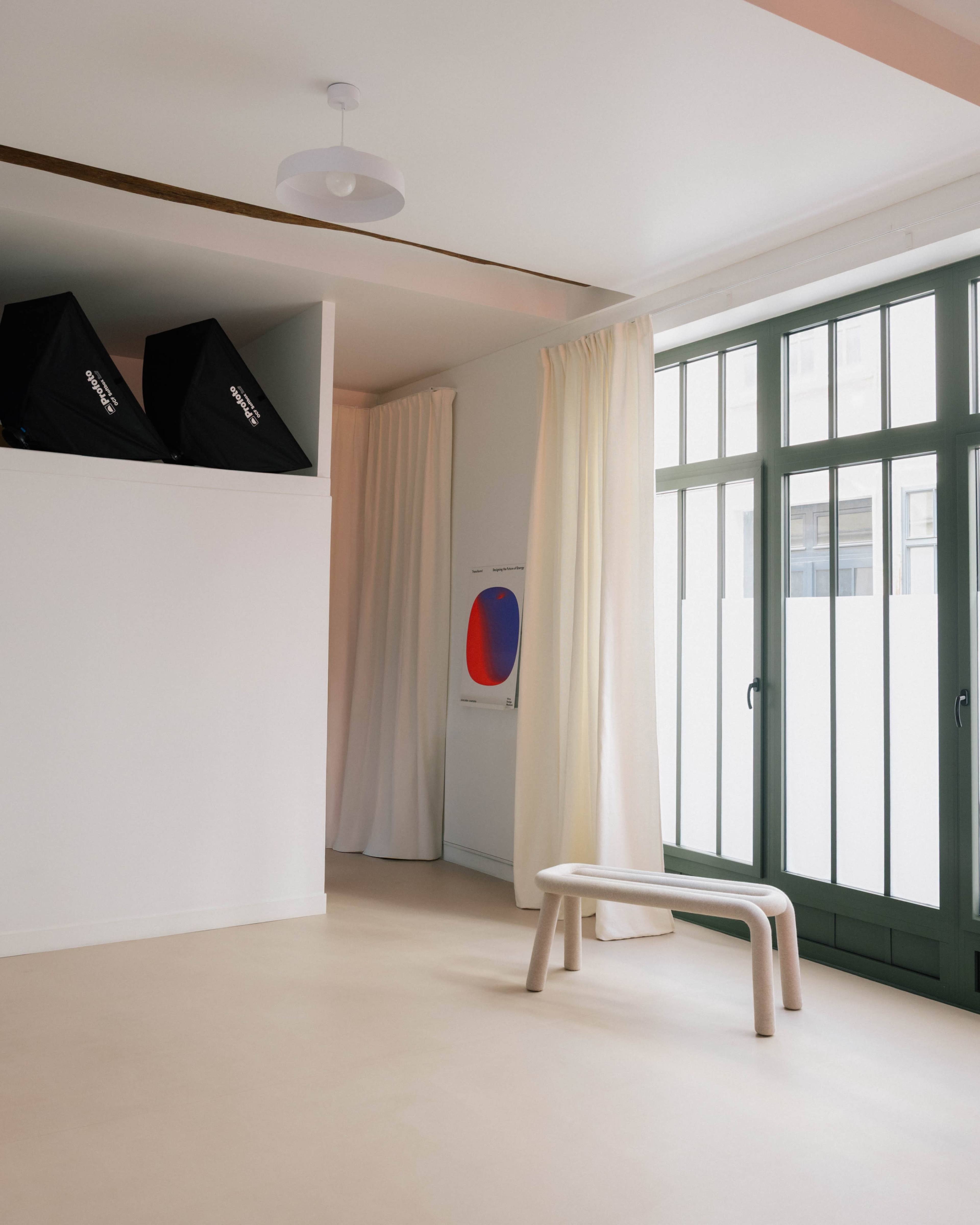 A bright room features a bench, two curtains, large windows, and photography equipment in the corner.