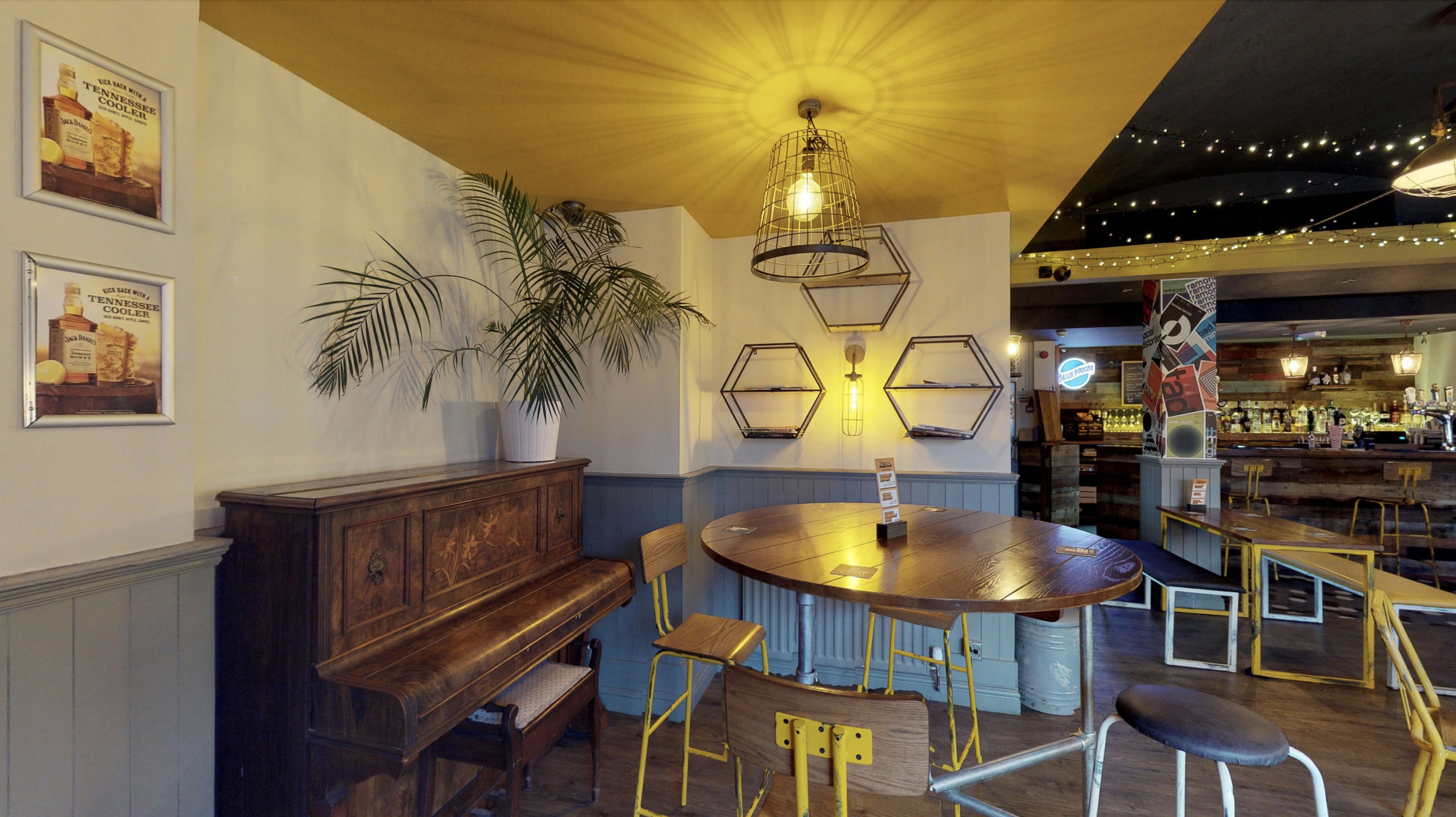 A cozy pub interior featuring a wooden piano, a round table with metal chairs, and decorative lighting fixtures on the walls.