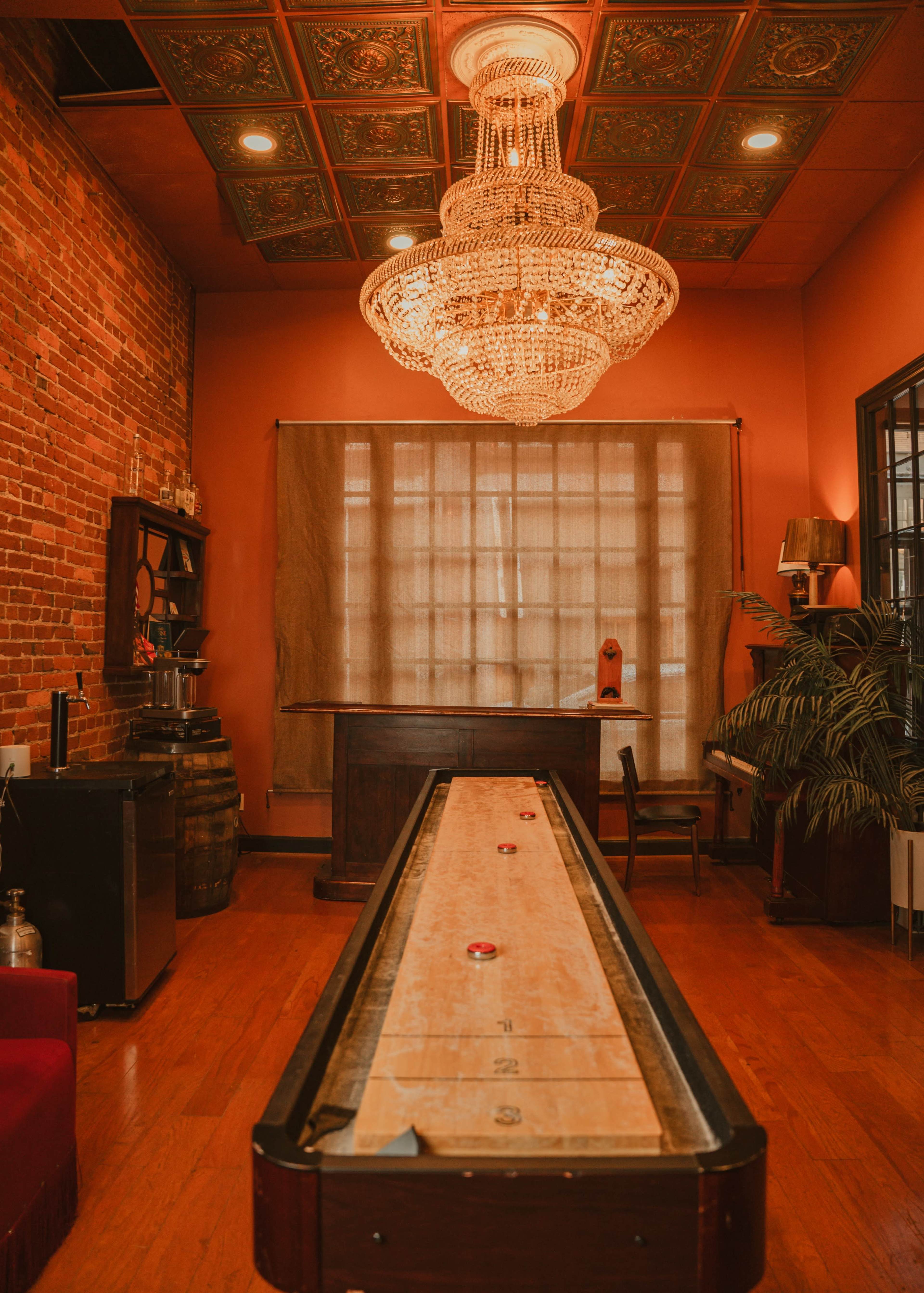 The interior features a long shuffleboard table in a warmly lit space with exposed brick walls and a large crystal chandelier.