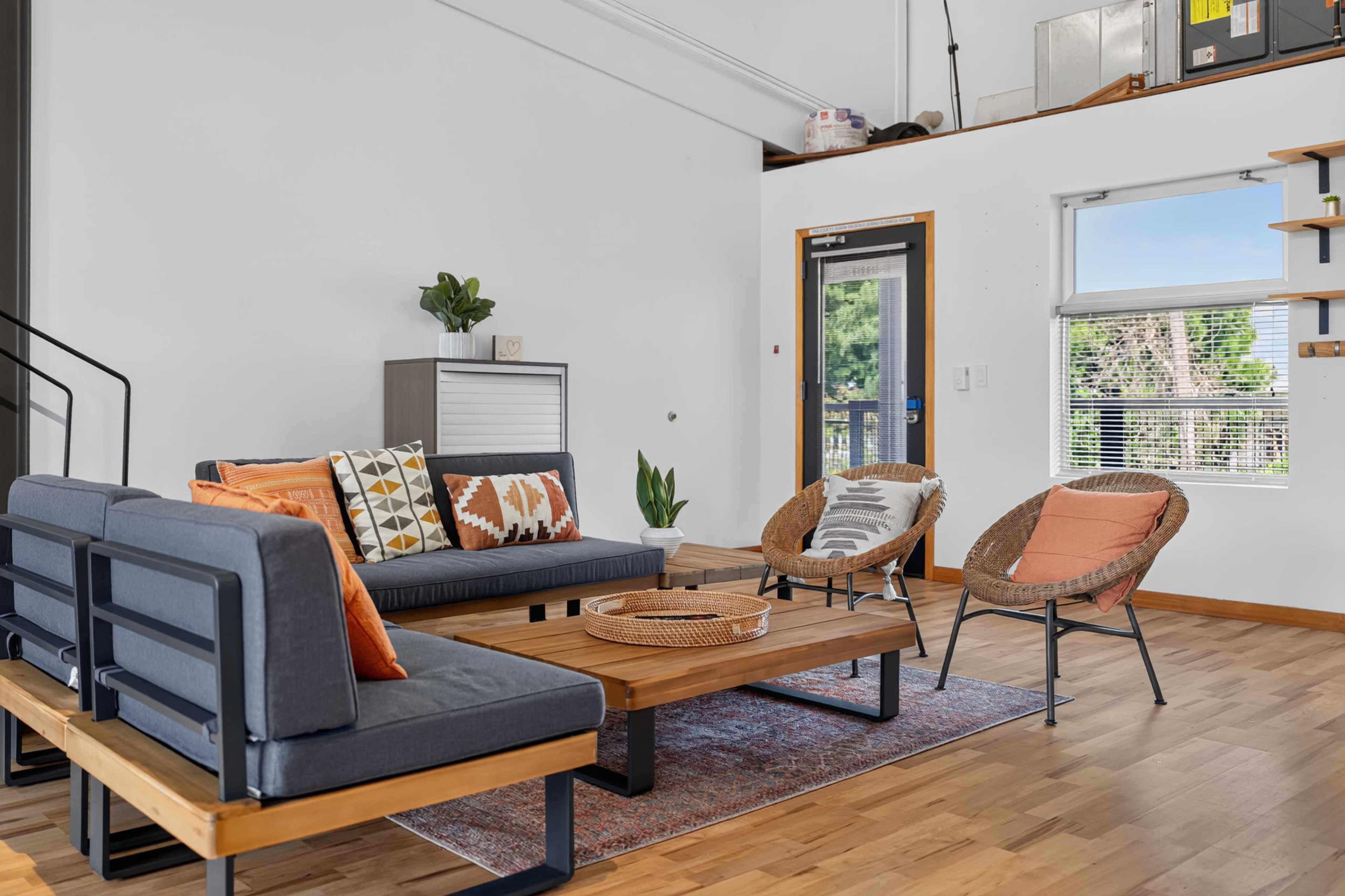 A modern living room features a seating area with a gray sofa and two rattan chairs, centered around a wooden coffee table on a patterned rug, with natural light streaming through a window.