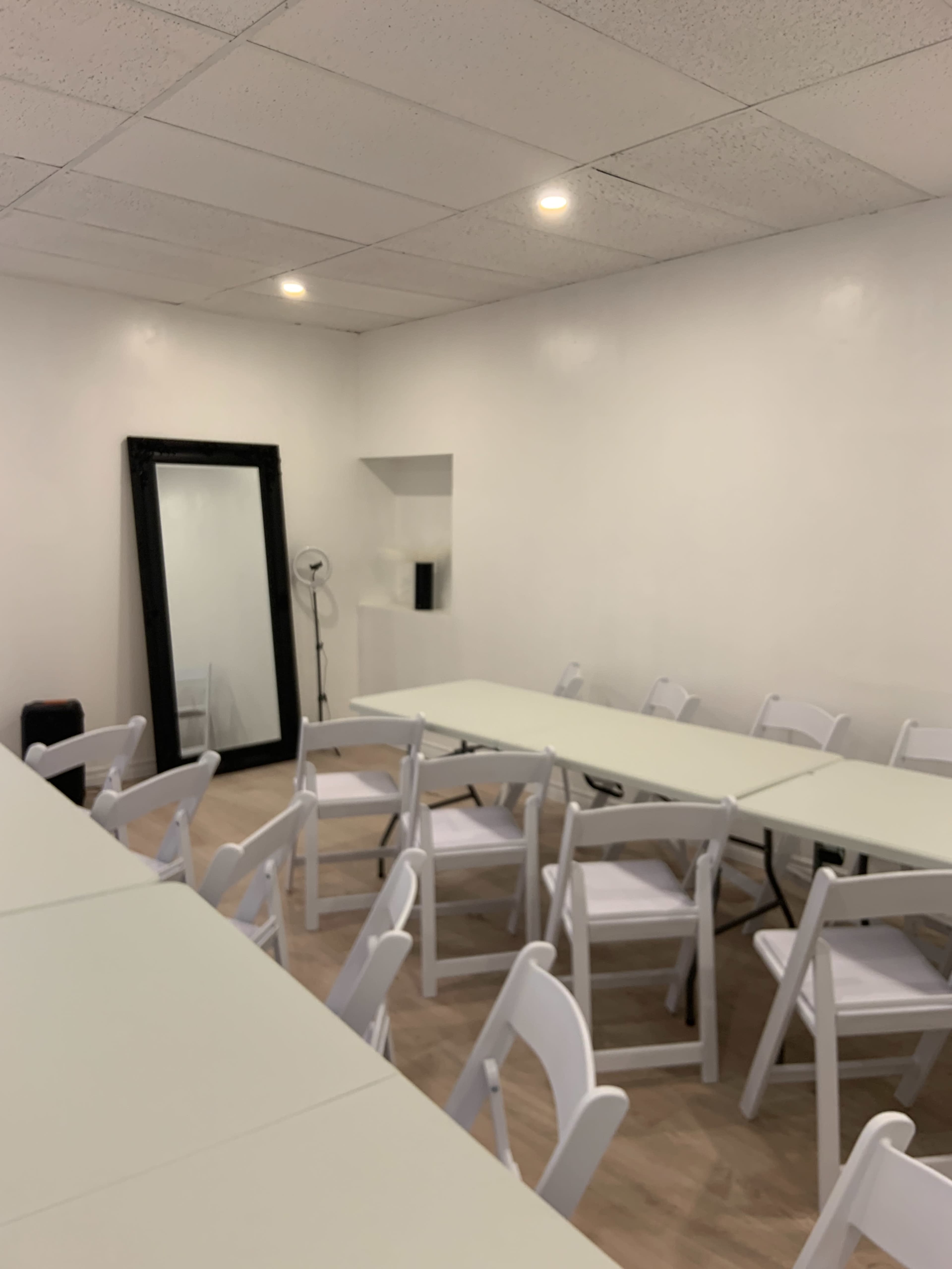 A sparsely furnished room with several tables arranged in a row and white folding chairs set around them, along with a large mirror on one wall.