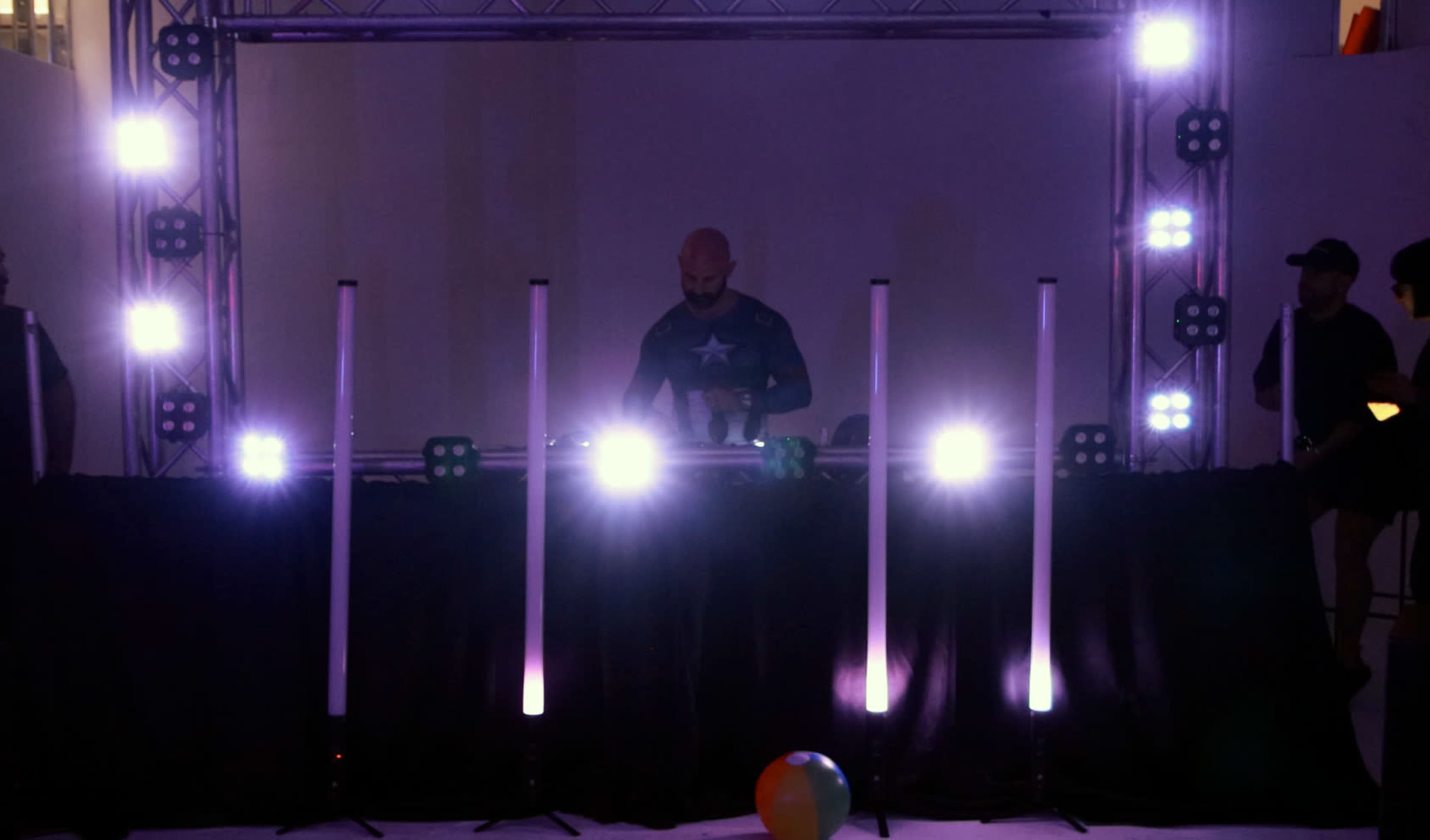 A DJ stands behind a console with illuminated vertical lights and a large crowd in the background.