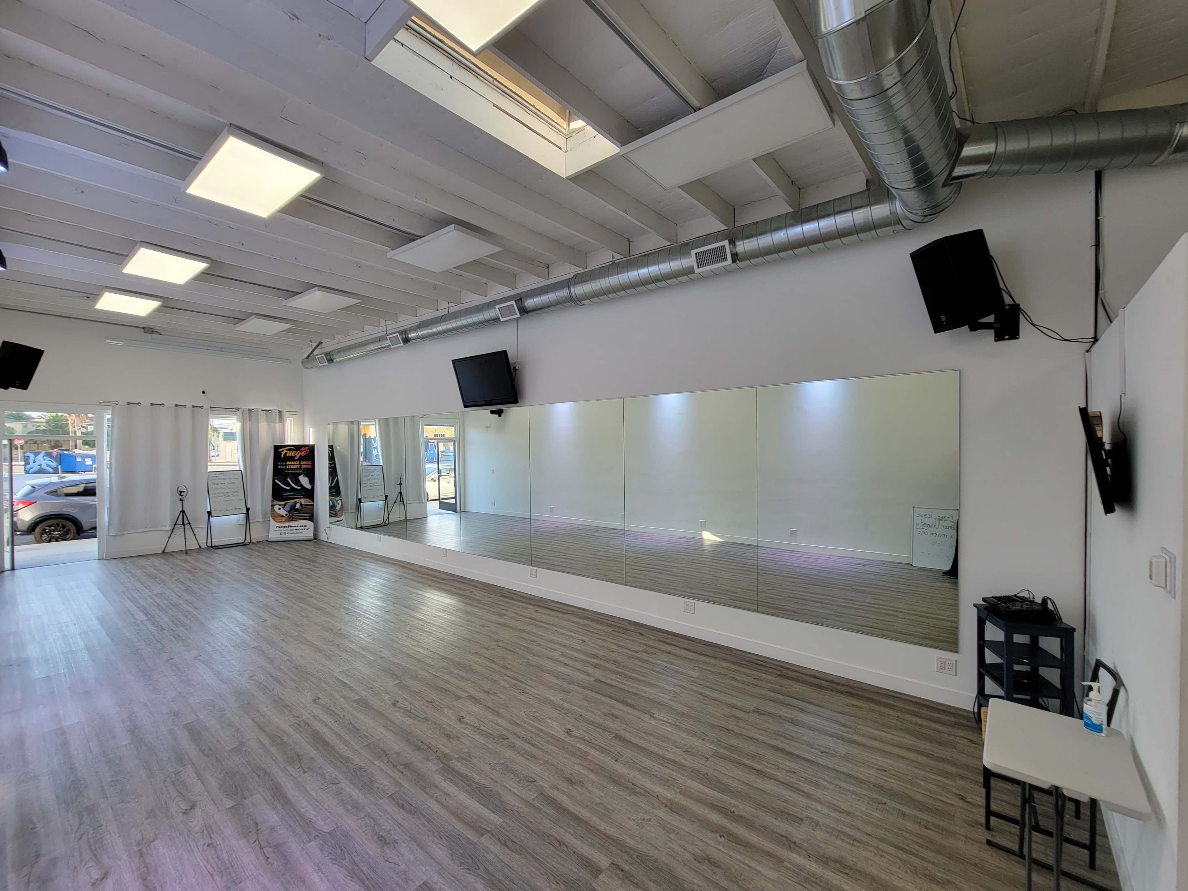 The image shows a spacious dance studio with wooden flooring, large mirrors on one wall, and natural light streaming in through a window.