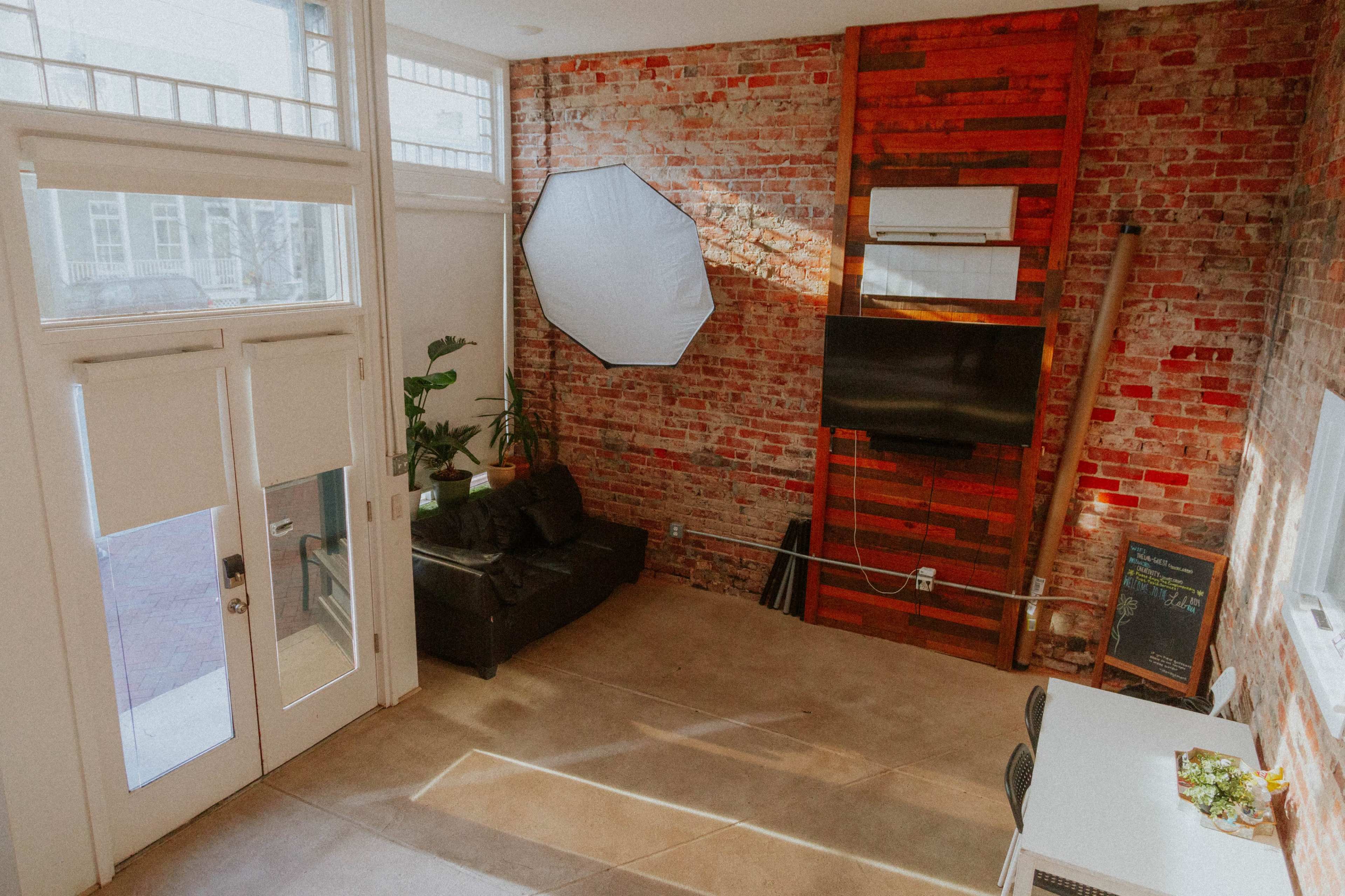 The image shows a modern interior space with exposed brick walls, a black sofa, a wall-mounted TV, and a small table with plants near large windows.