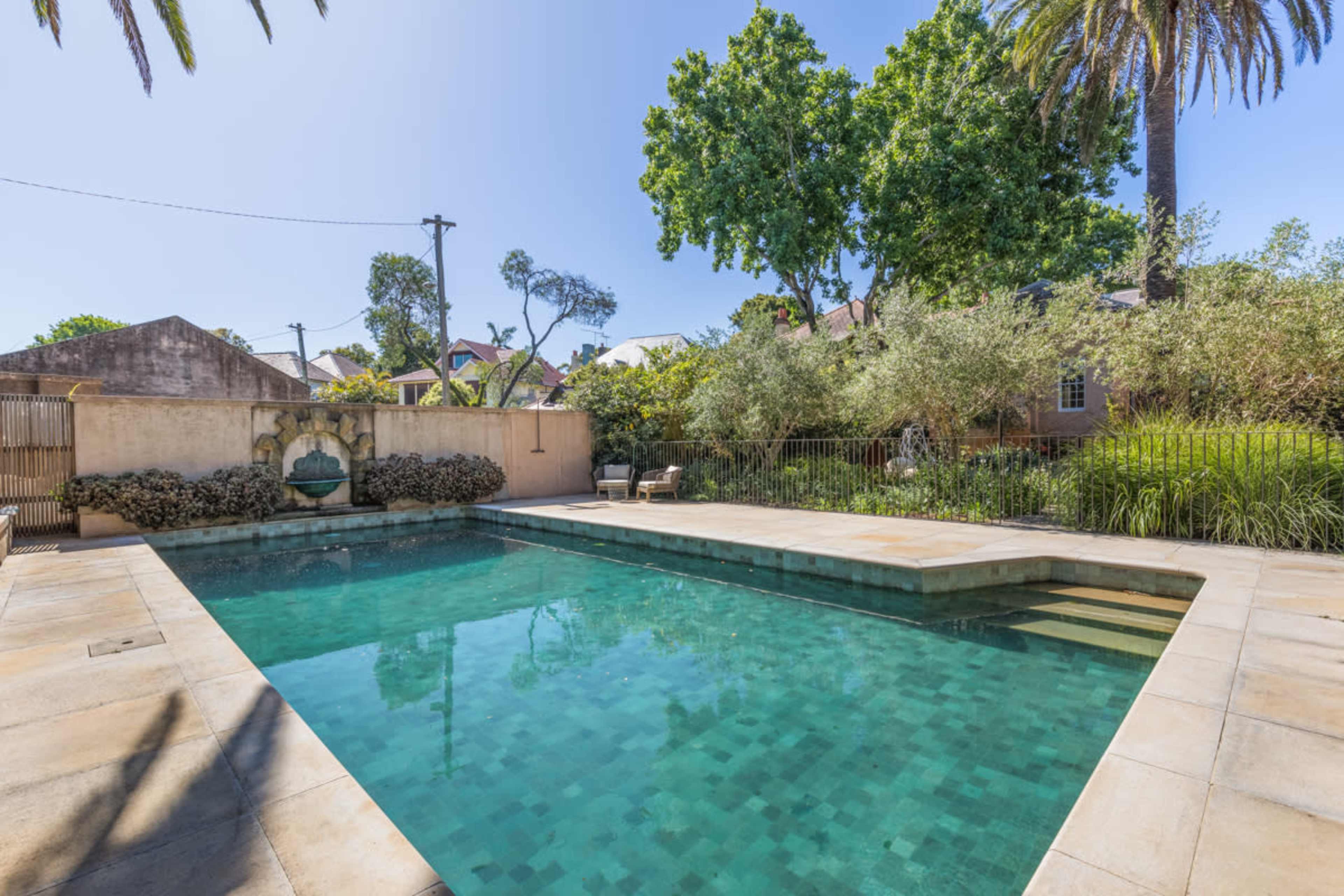 The image shows a swimming pool surrounded by lush greenery and palm trees, with a small seating area visible nearby.