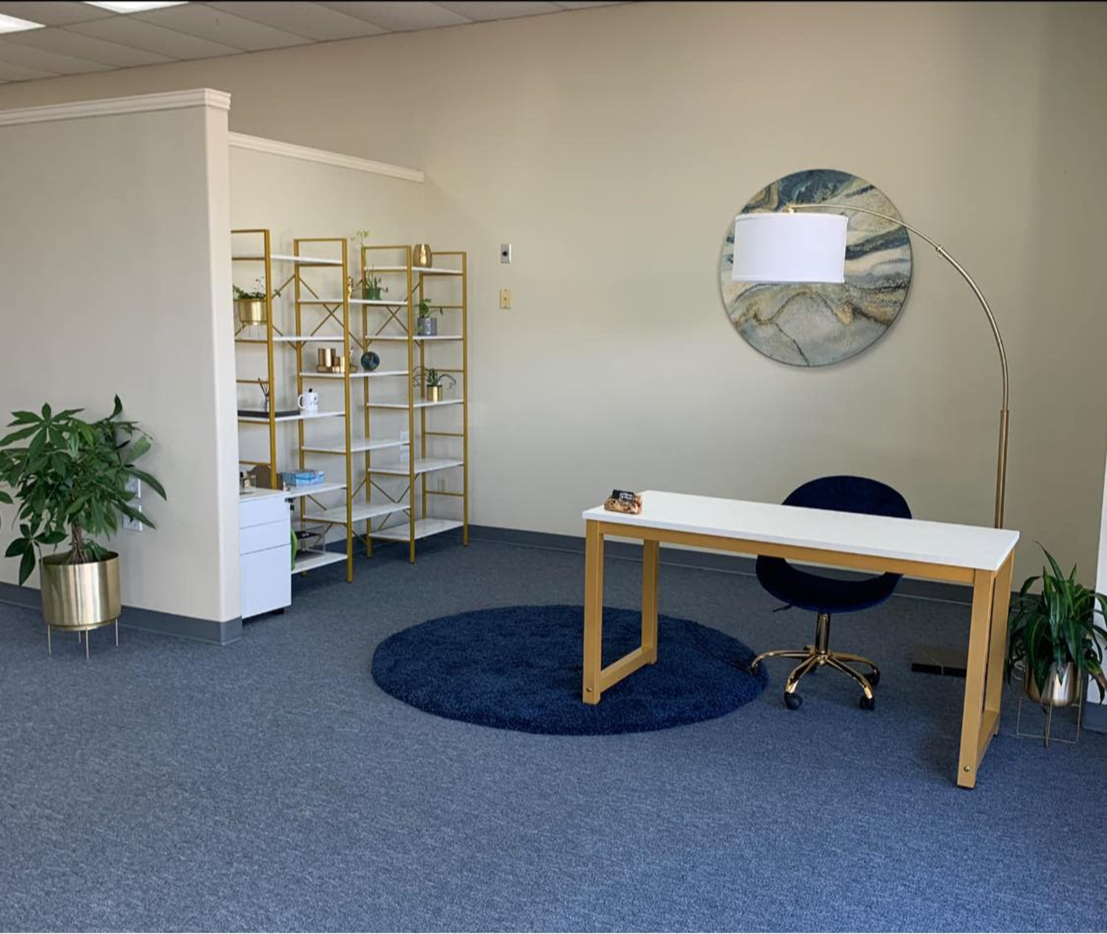The image shows a modern office space featuring a white desk with a dark blue chair, a round rug, a bookshelf with decorative items, and a floor lamp.