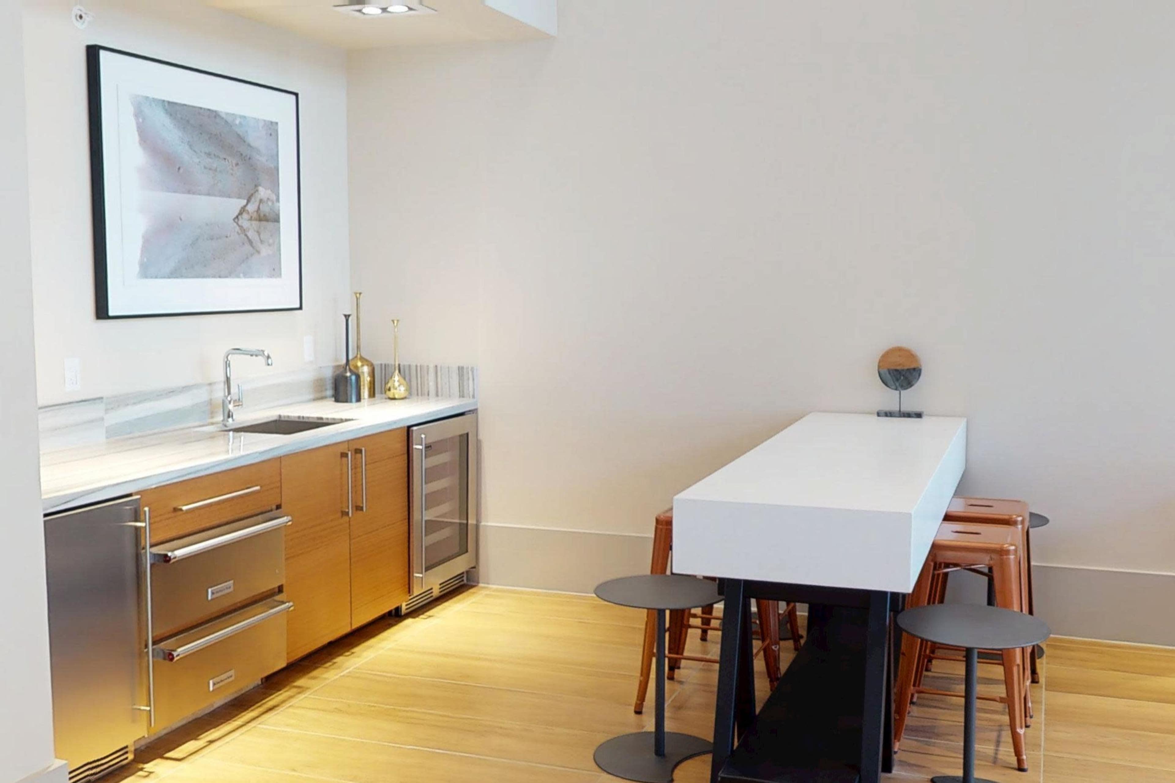 The image depicts a modern kitchen with a wooden cabinetry, stainless steel appliances, and a white dining table accompanied by several stools.