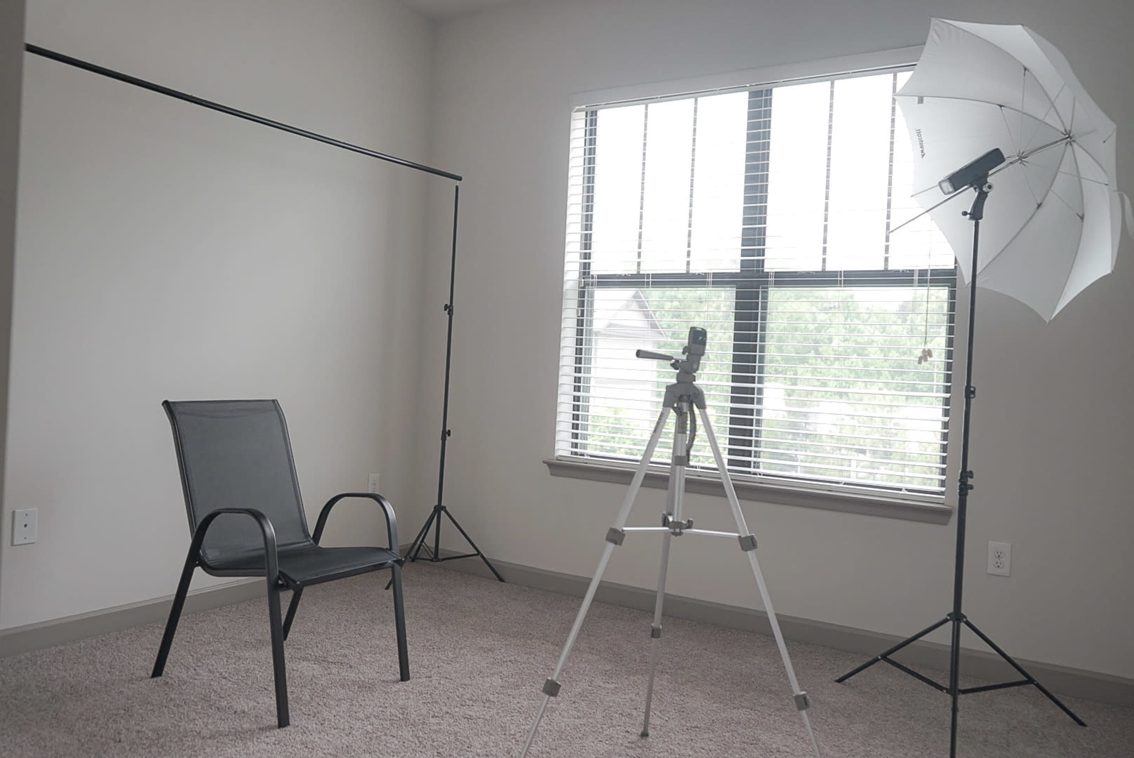 A bare room with a chair, a light stand with an umbrella, and a tripod positioned near a window with blinds.