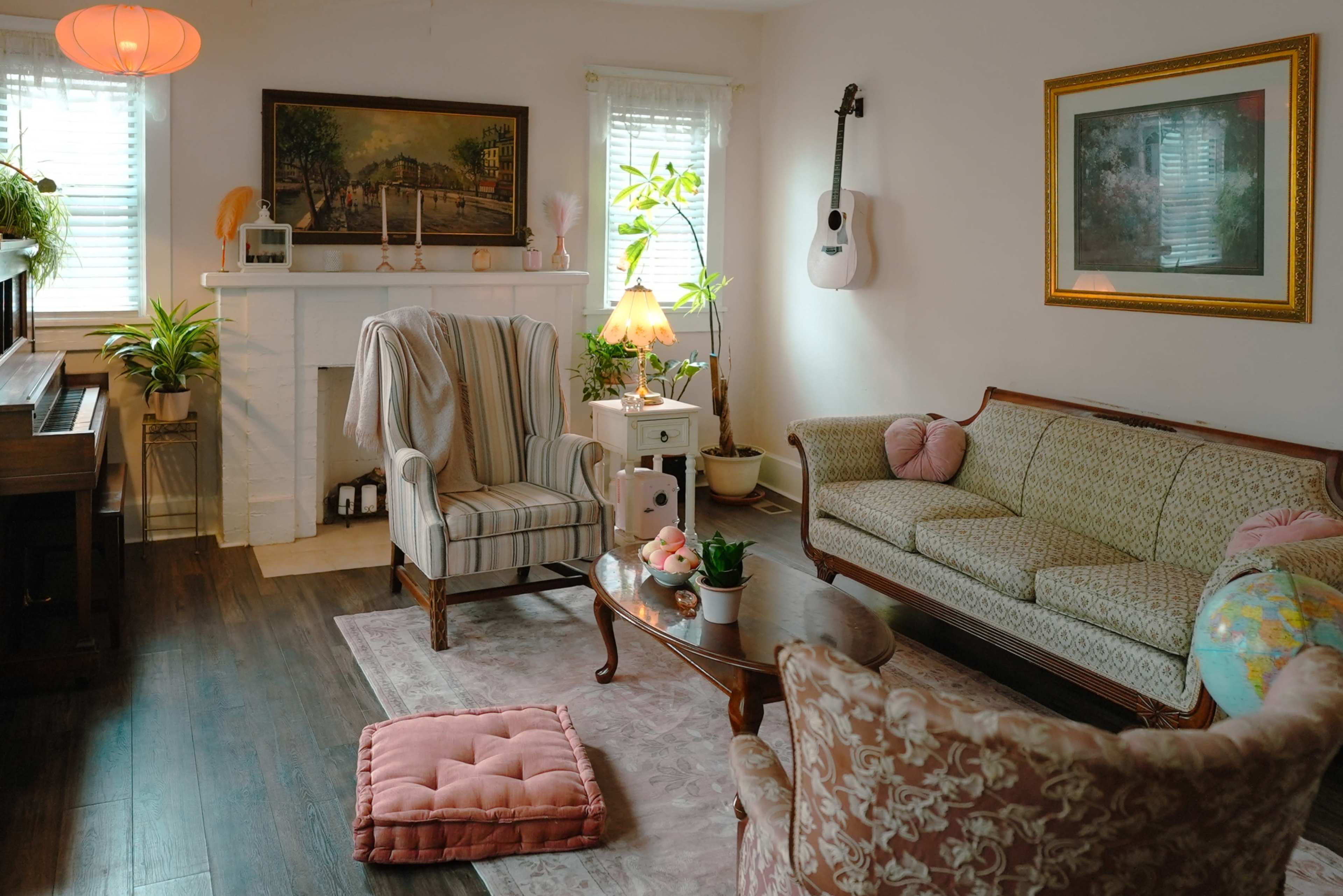 The image shows a cozy living room with a mix of vintage furniture, including a patterned couch and armchairs, a wooden coffee table, and a fireplace adorned with plants and decor.