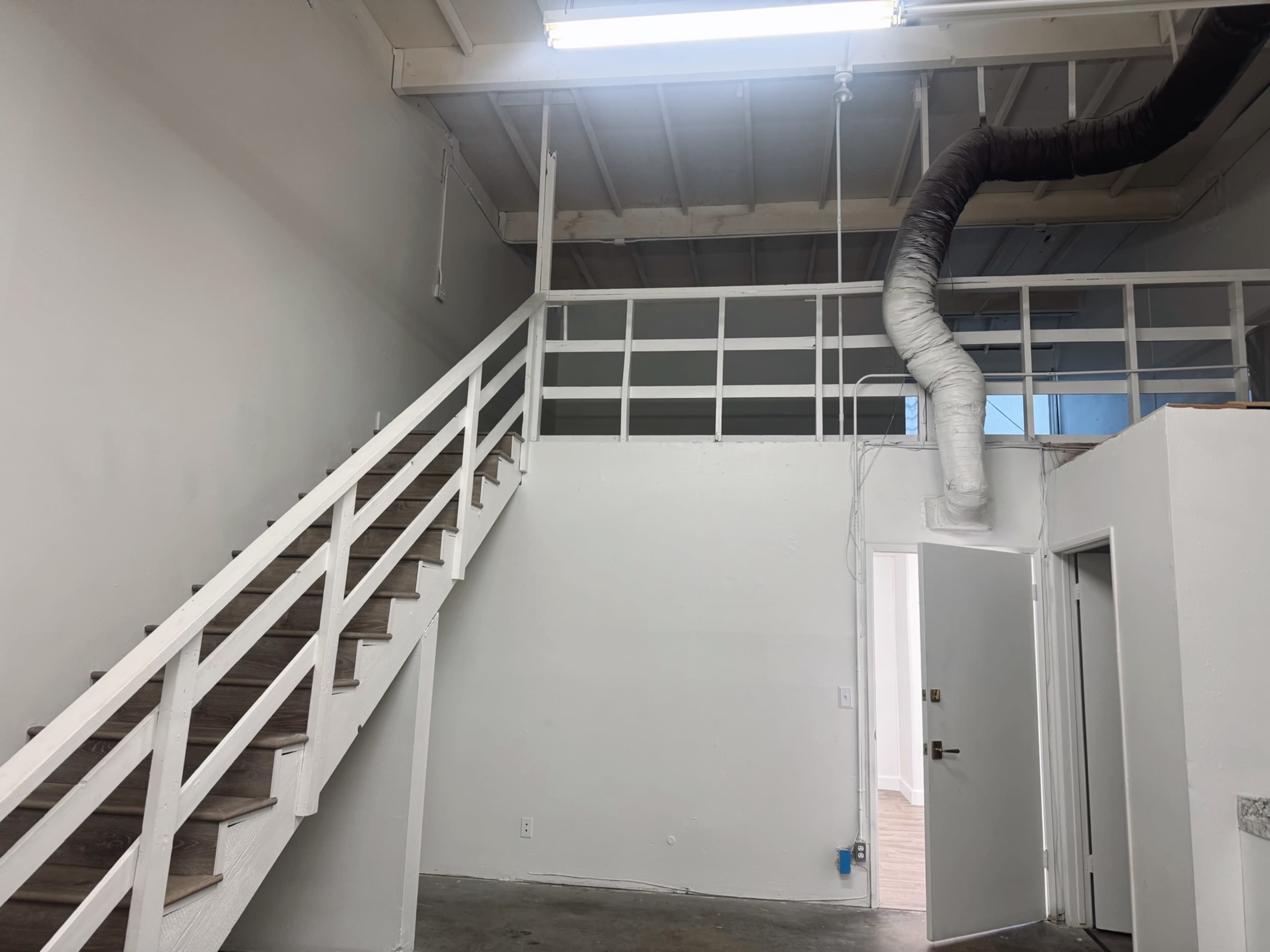 The image shows a interior space featuring a staircase leading to a loft area, with white walls and a visible ventilation duct running along the ceiling.