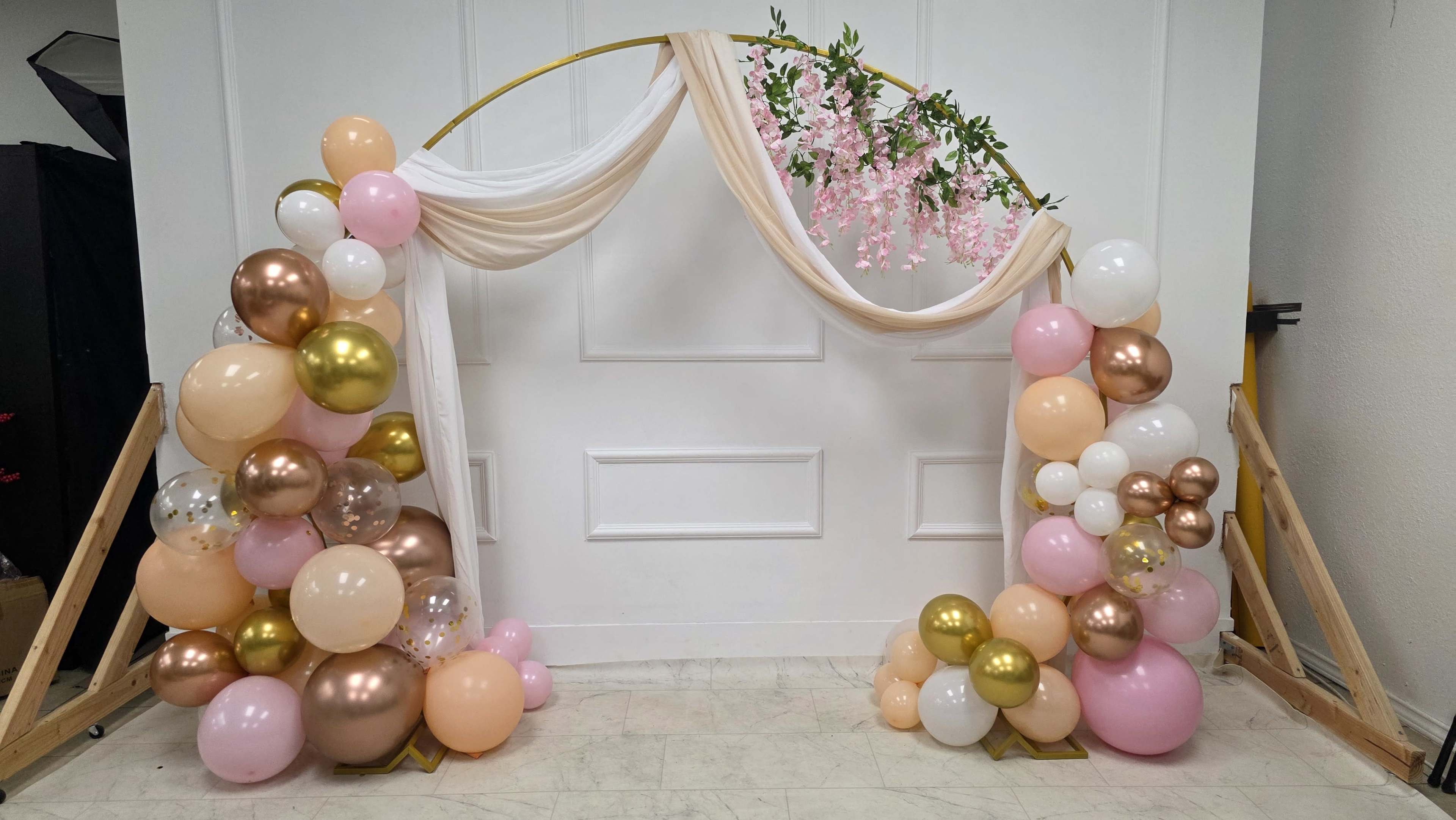 The image features a decorative arch adorned with draped fabric and artificial flowers, flanked by clusters of balloons in various colors and sizes.