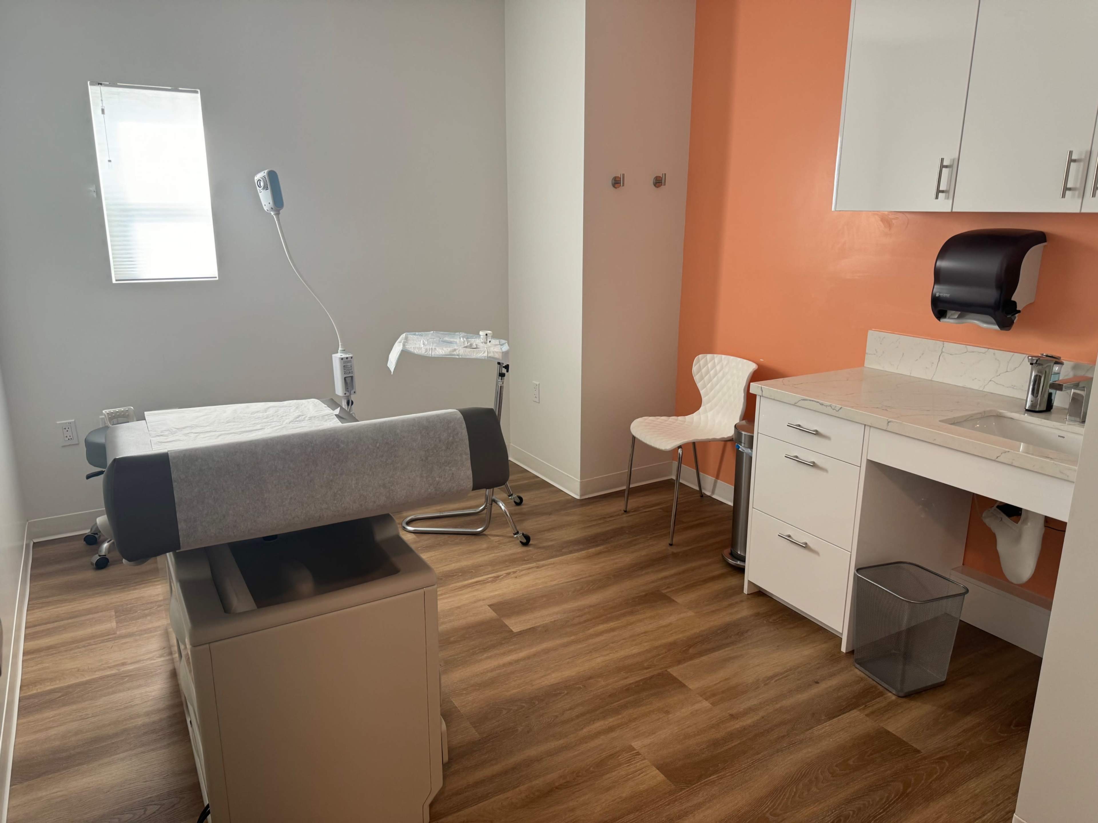 A modern medical examination room with a treatment table, a chair, and a sink area against an orange accent wall.
